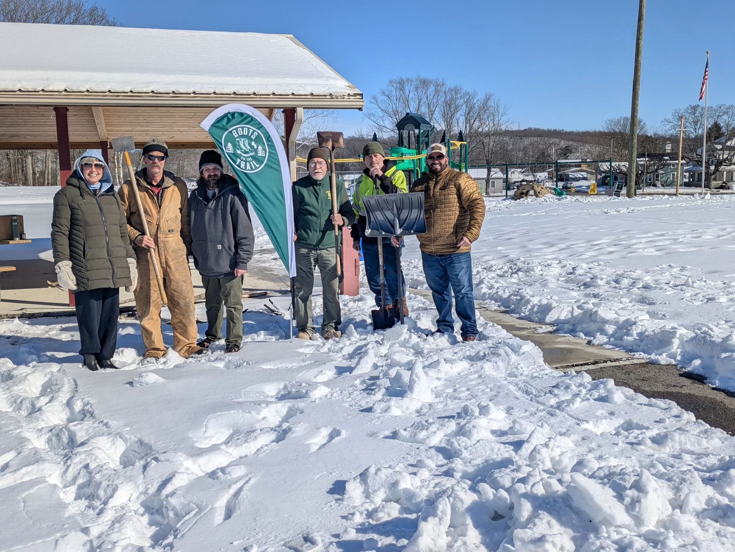 Group of people standing in snow outside near a pavilion, some holding shovels, with a banner that reads 'Boots on the Trail', a playground and American flag in the background.