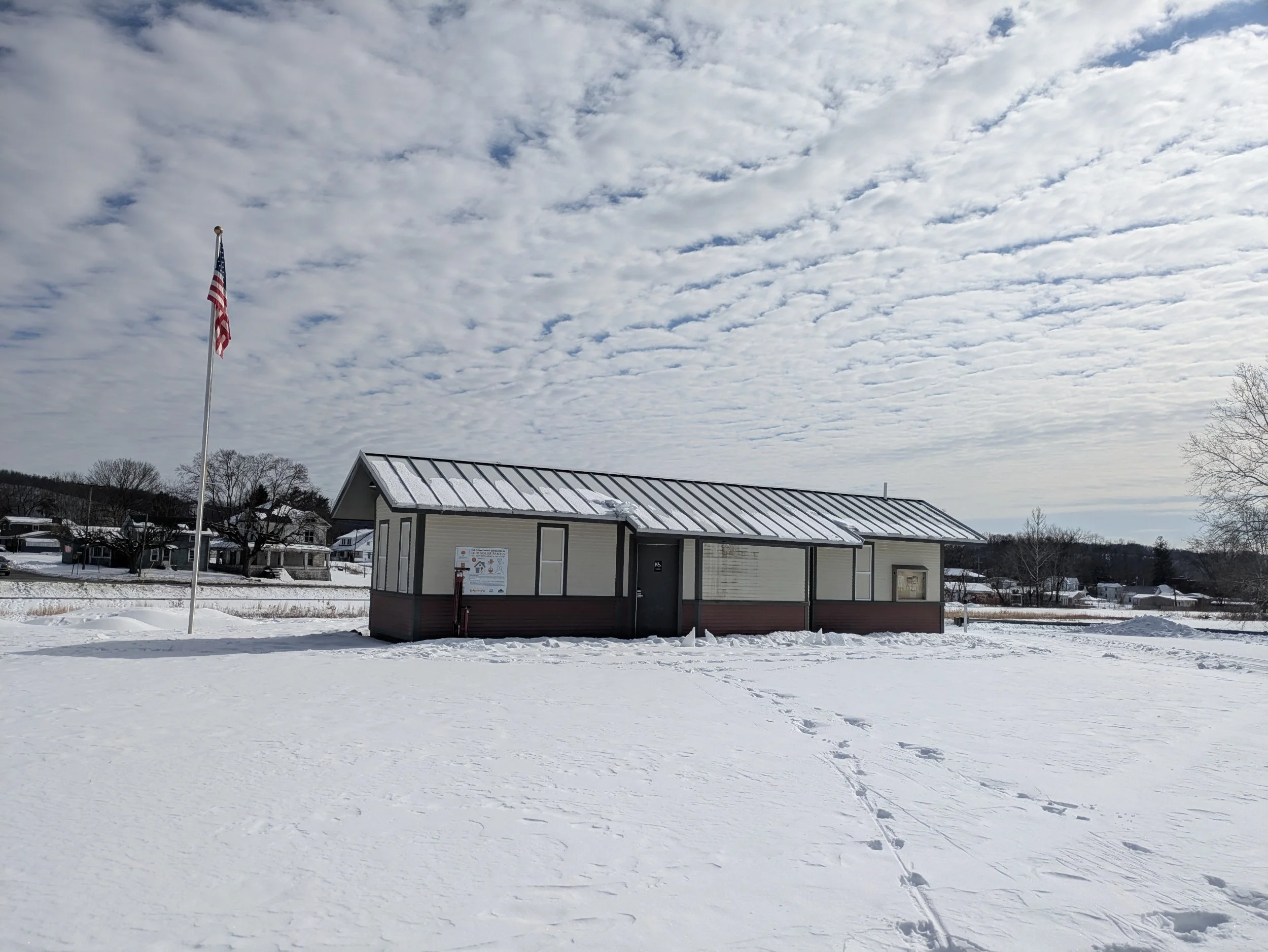 A small cream-colored building with a metal roof and bulletin boards on the exterior, situated in a snowy landscape under a cloudy sky, with an American flag on a flagpole nearby.