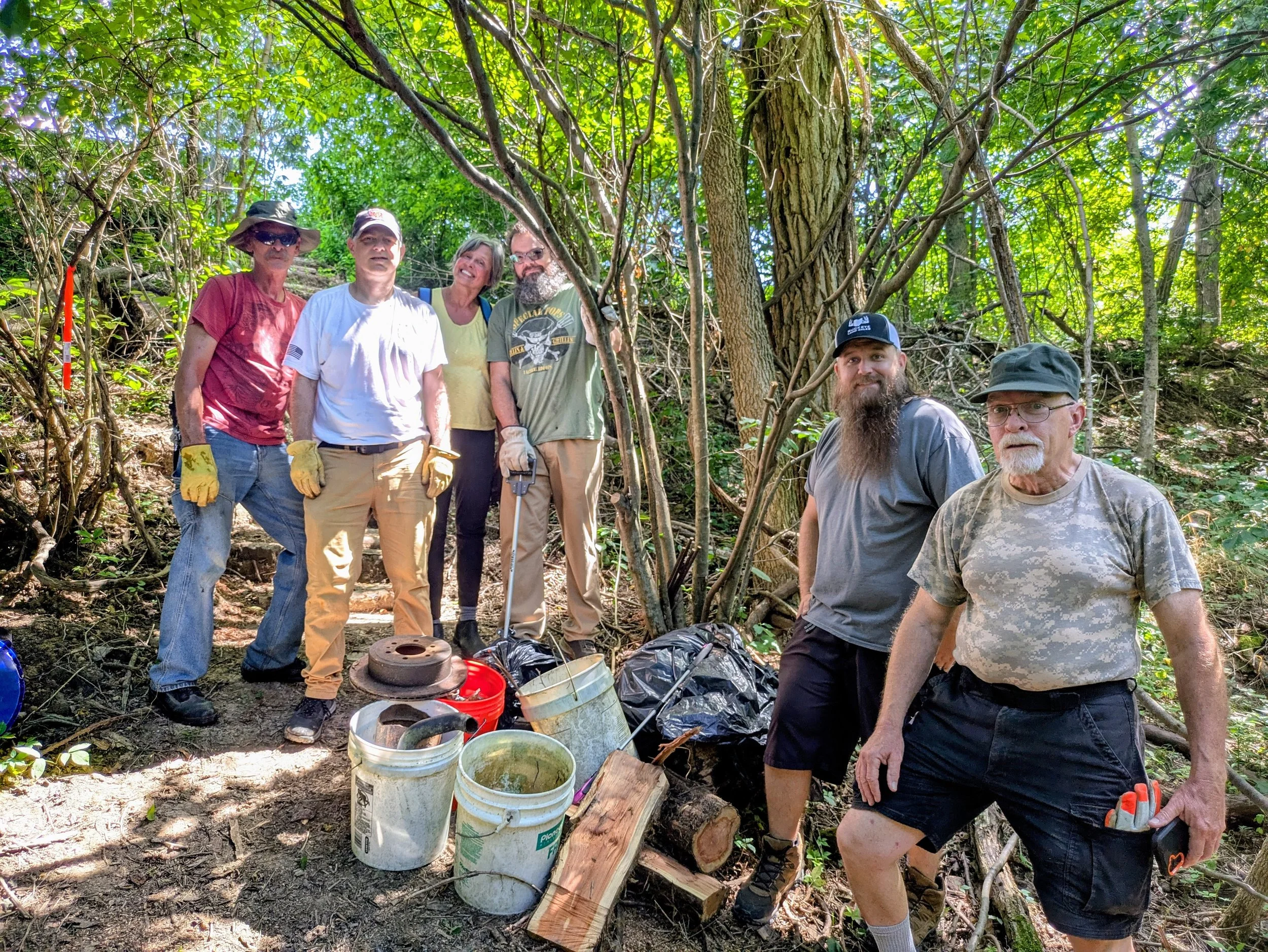 A group of seven people in a wooded outdoor setting during daytime, some wearing work gloves and hats, standing near buckets and collected debris, engaged in a cleanup or conservation activity.
