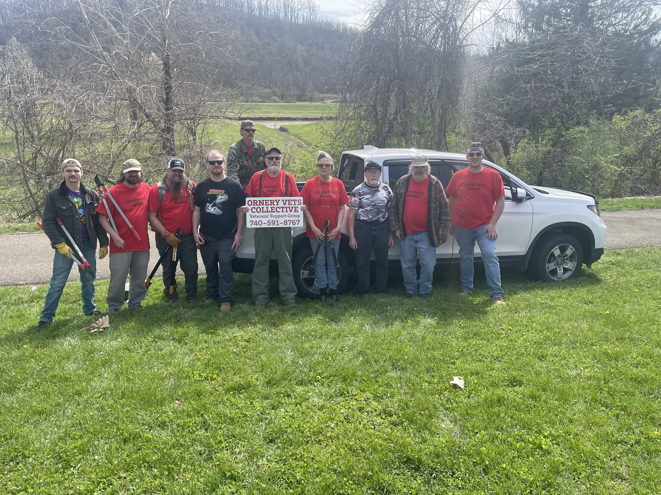A group of people standing outdoors, some holding tools, posing behind a white vehicle with a sign that reads "ORNERY VETS COLLECTIVE Veterans Support Group 740-591-8767". They are dressed casually and are surrounded by trees and grassy area.