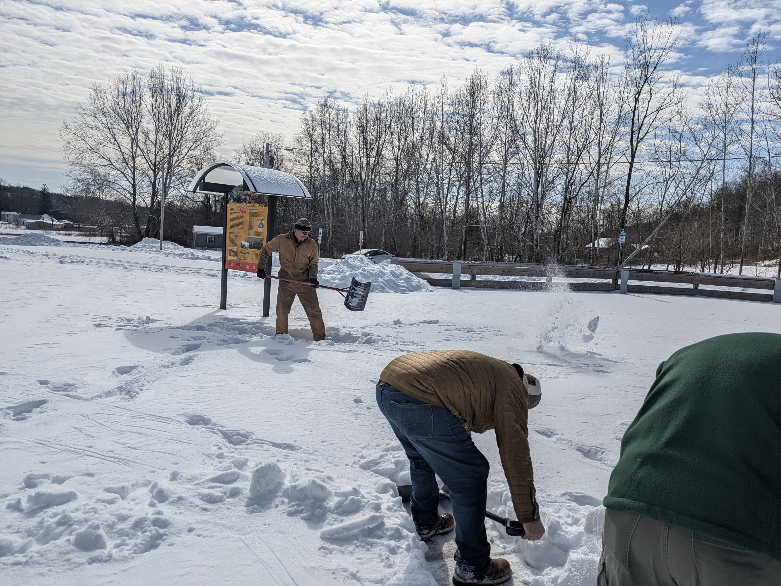 Two men shoveling snow in an open park with snow-covered ground, leafless trees, and a cloudy sky.