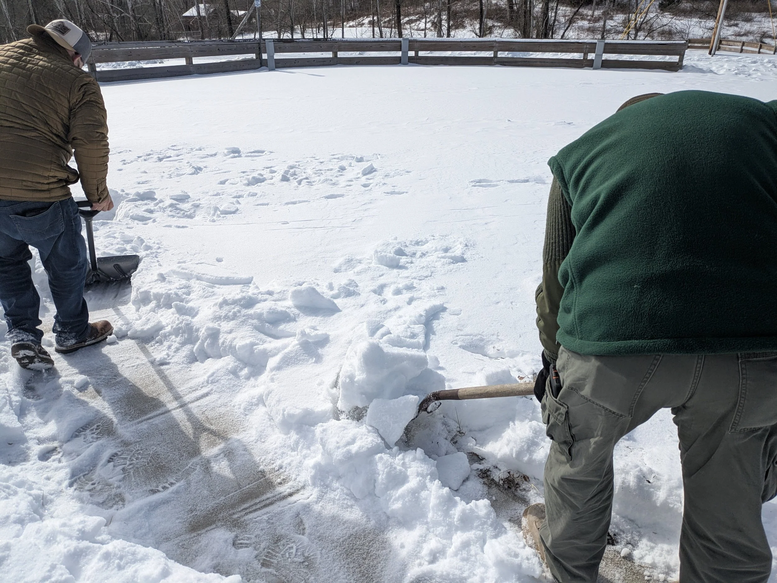 Two people are shoveling snow on a deck or patio area, with snow-covered ground and a wooden fence in the background. One person is on the left, wearing a brown jacket and a gray hat, and the other on the right, wearing a green vest and khaki pants.