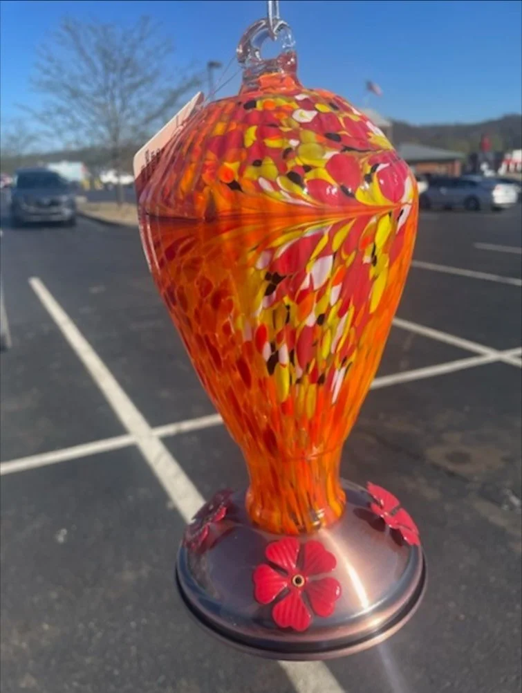 Colorful glass hummingbird ornament with red flower accents, hanging in an outdoor parking lot against a blue sky.