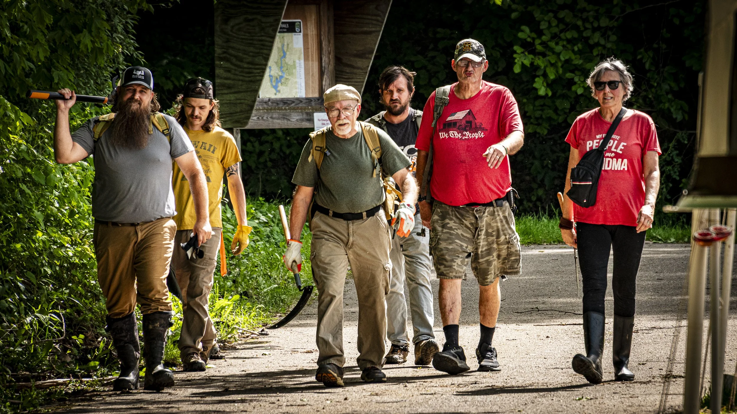 Group of six hikers walking along a trail surrounded by trees, wearing casual outdoor clothing, backpacks, and holding hiking gear.