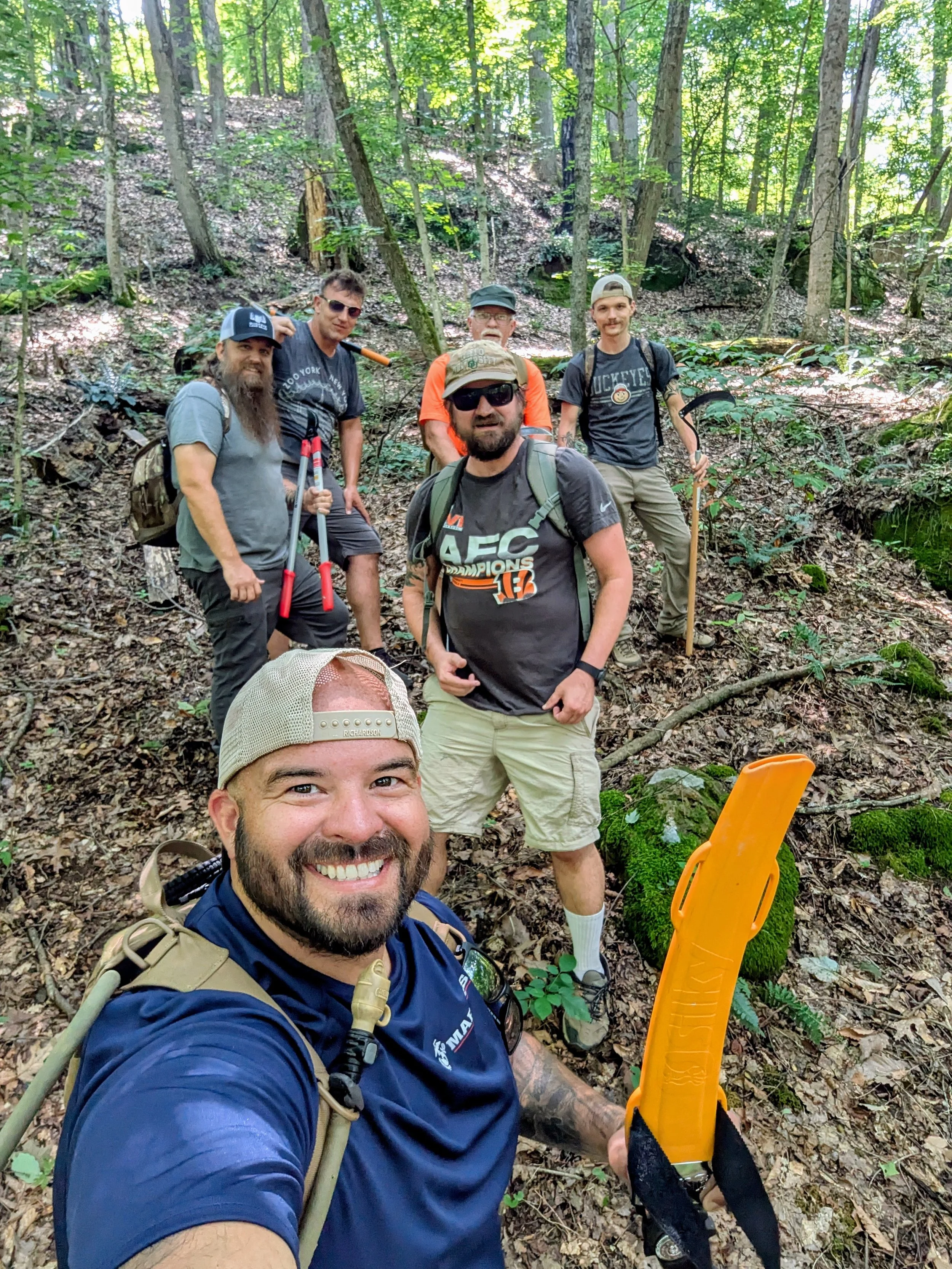Five men in shorts and t-shirts outdoors in a wooded area, taking a selfie during a hike or outdoor activity. One man holds a yellow plastic object, and others have backpacks and hiking gear.