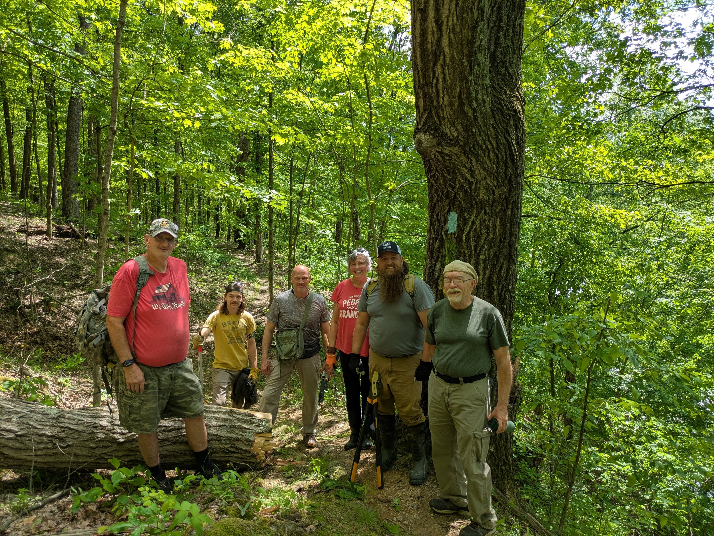 A group of eight hikers standing on a trail in a dense green forest, with tall trees and leafy canopy, some smiling at the camera.