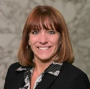 Woman with shoulder-length red hair, wearing a black blazer with a patterned shirt, smiling, against a gray background.