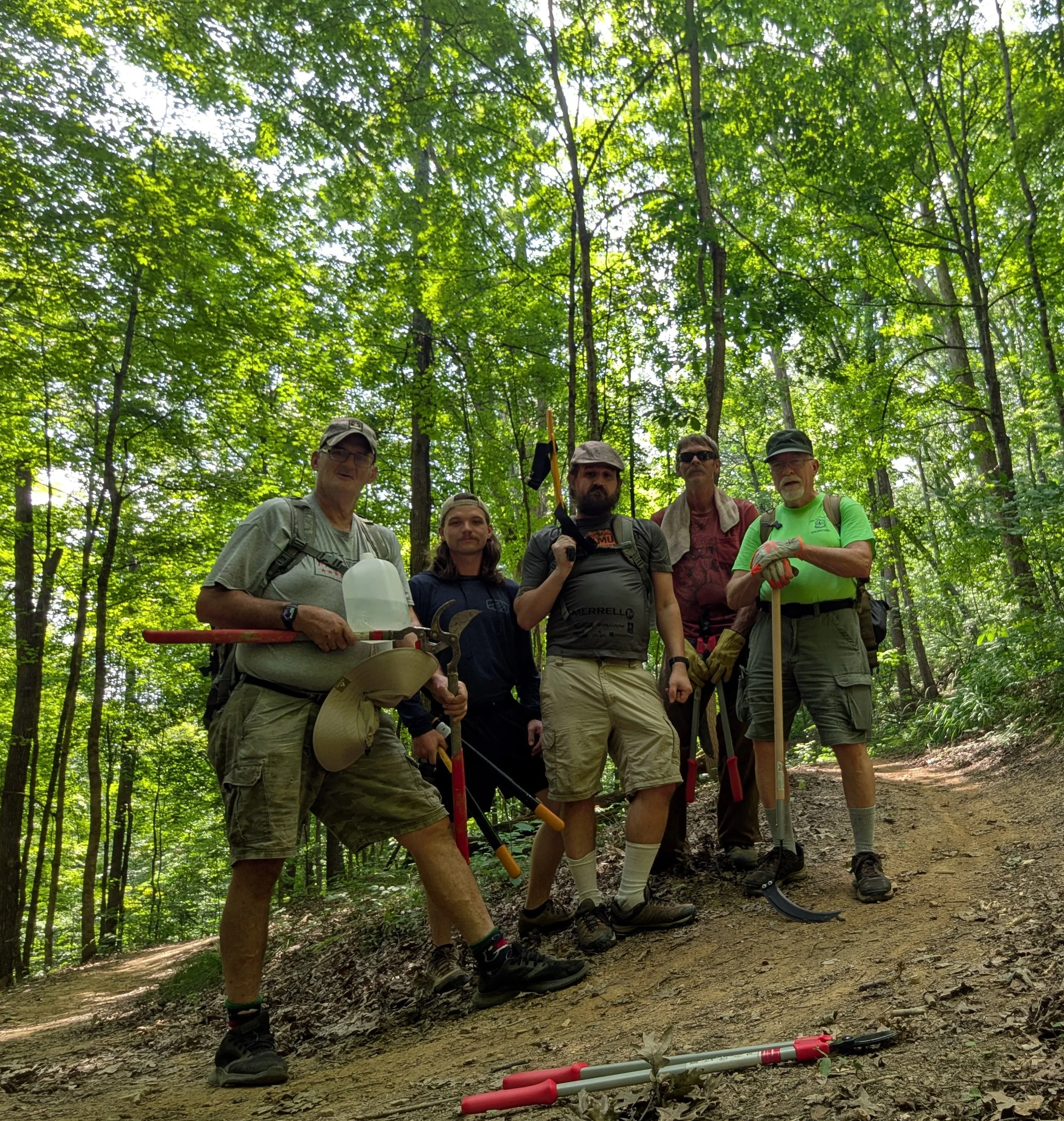 A group of six men standing on a dirt trail in a dense, green forest, wearing outdoor or hiking gear, with some holding walking sticks or tools, and trees with lush foliage overhead.