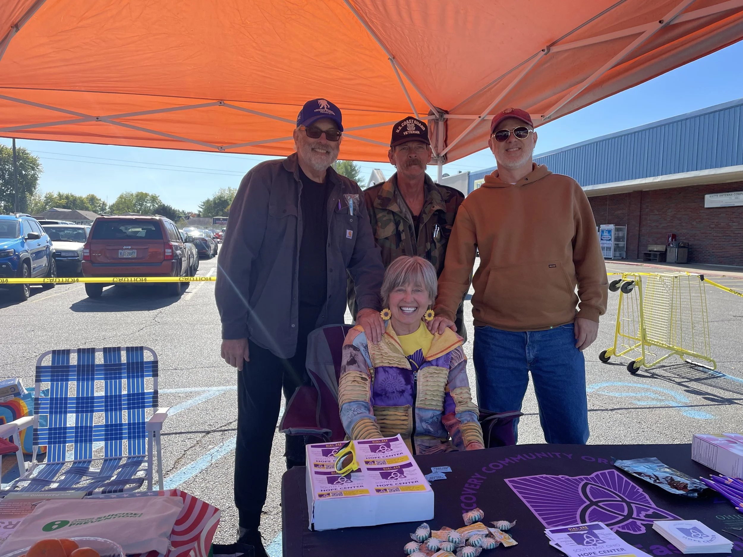 Group of four people, three men and one woman, standing under an orange canopy, smiling at the camera. The woman is seated and holding sunflower earrings. The men are wearing casual clothing, with two of them wearing hats and sunglasses. There are parking lot and store buildings in the background, along with tables and promotional materials on the table in the foreground, indicating an outdoor community event or outreach.