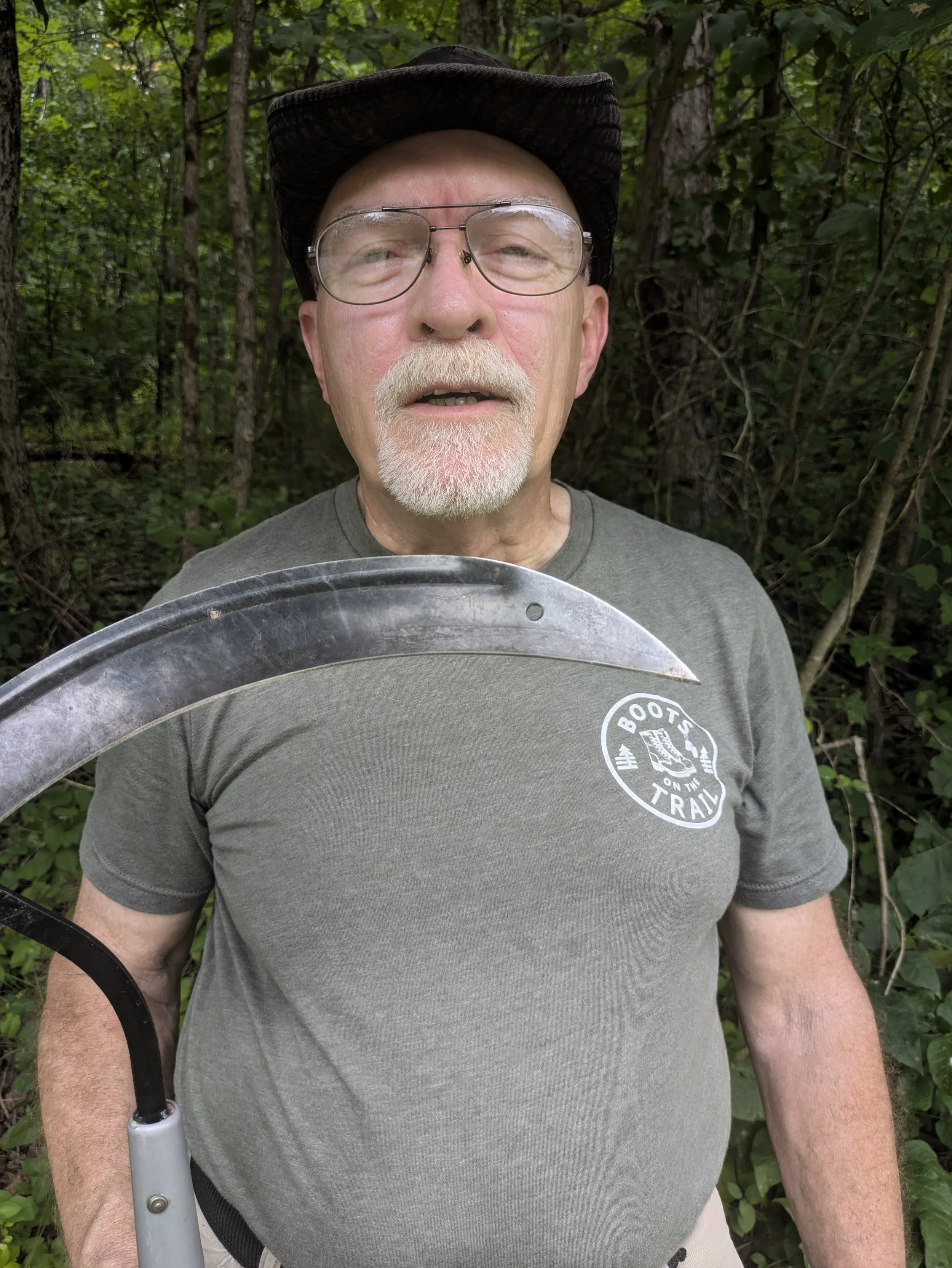 An elderly man with glasses, a gray beard, and a yellow shirt is pruning bushes with hedge clippers in a dense shrubbery. He is wearing a beige cap with a bicycle drawing on it and orange and white gardening gloves.