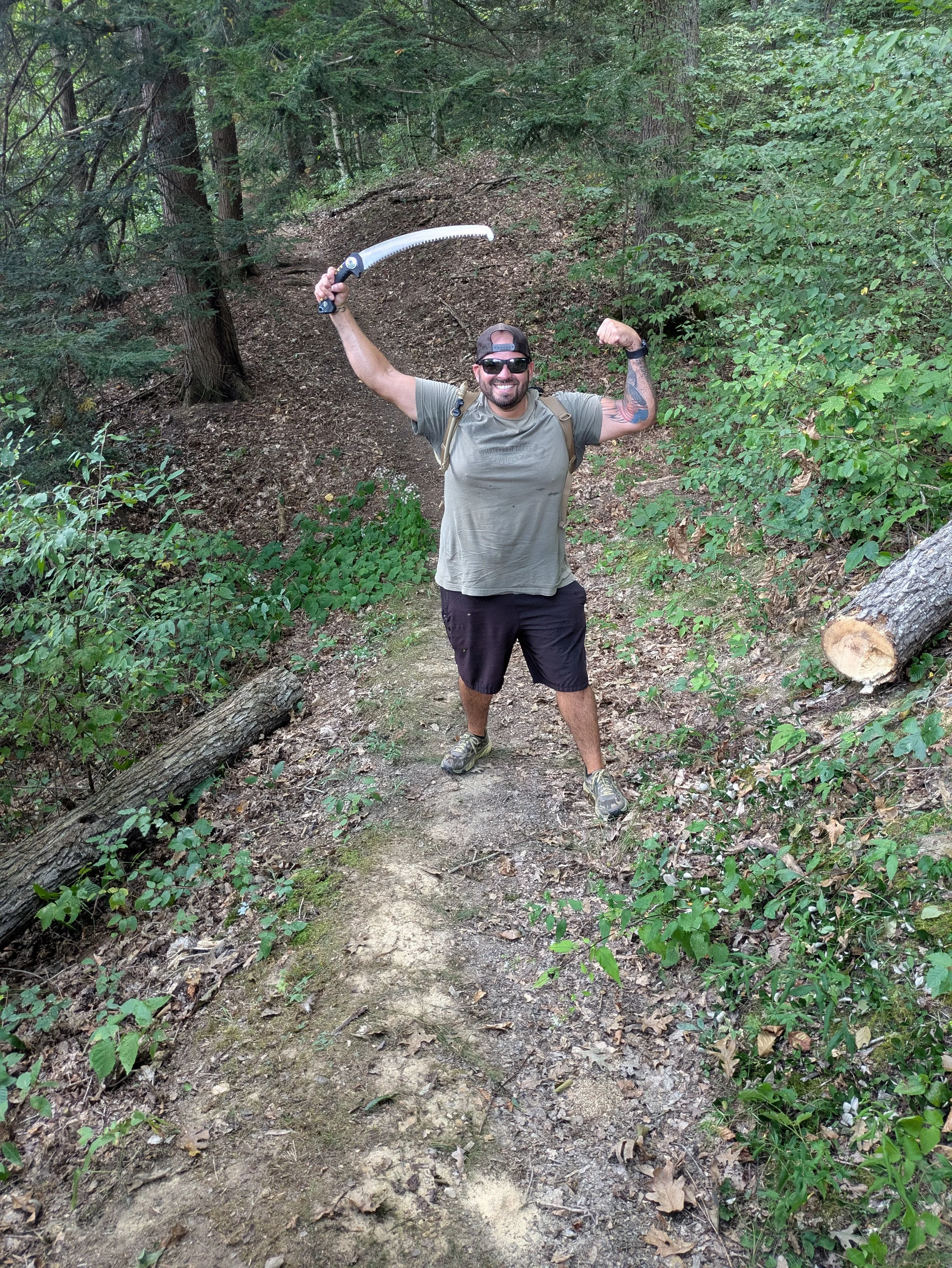 A man standing on a forest trail, holding a machete in his right hand and flexing his left arm. He is wearing sunglasses, a cap backward, a tan T-shirt, black shorts, and hiking shoes. The forest is dense with green foliage, trees, and fallen logs.