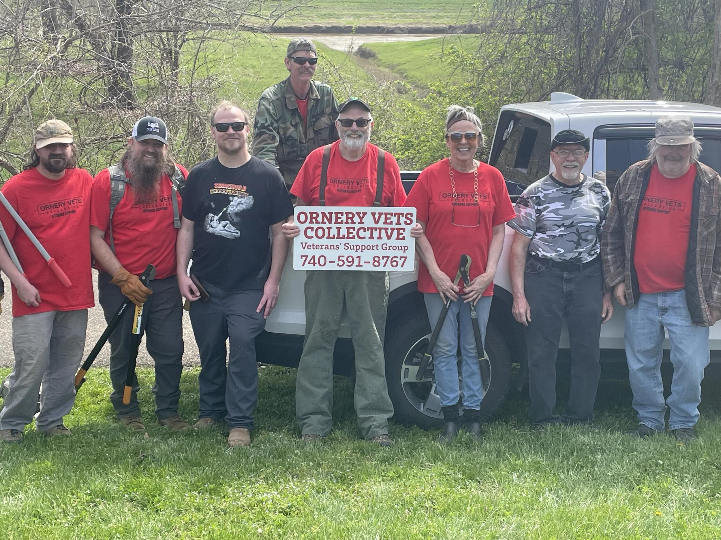A group of nine people standing outdoors in front of a pickup truck, with trees and a road in the background. Some are wearing red shirts with the text 'Ornery Vets Collective,' and one person holds a sign with the same text and a phone number, indicating they are part of a veterans support group.