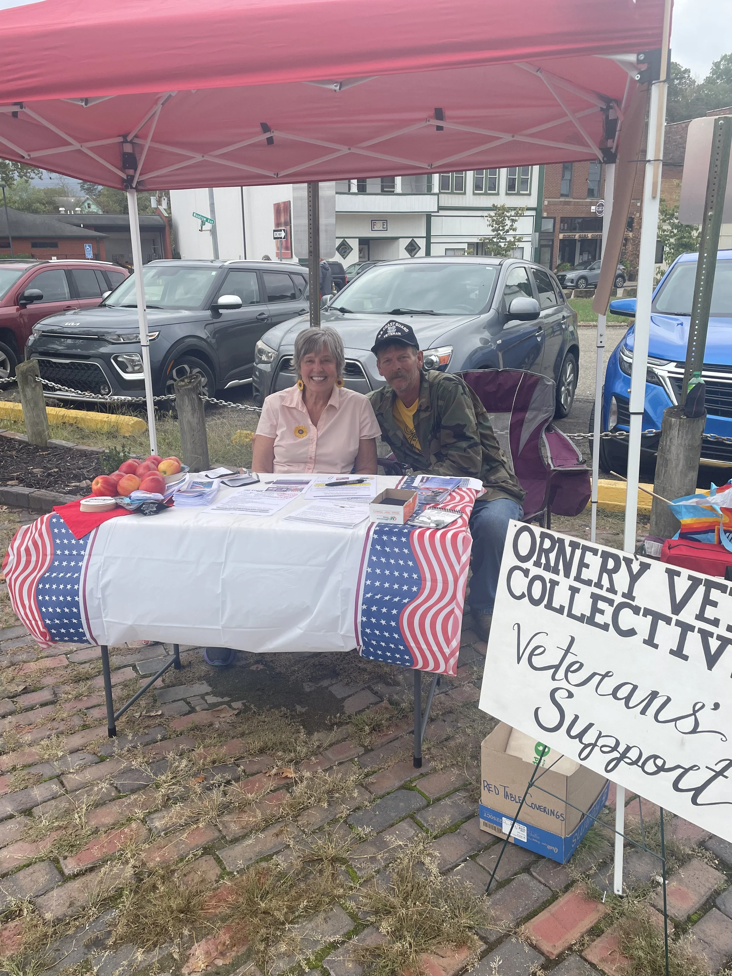 A woman and a man sitting behind a table at an outdoor event under a pink canopy. The table is covered with an American flag tablecloth and has peaches, papers, and other items on it. A sign in front of the table indicates it is part of the Orleans Vets Collectives' event, and the woman and man are smiling for the photo. There are parked cars and buildings in the background.
