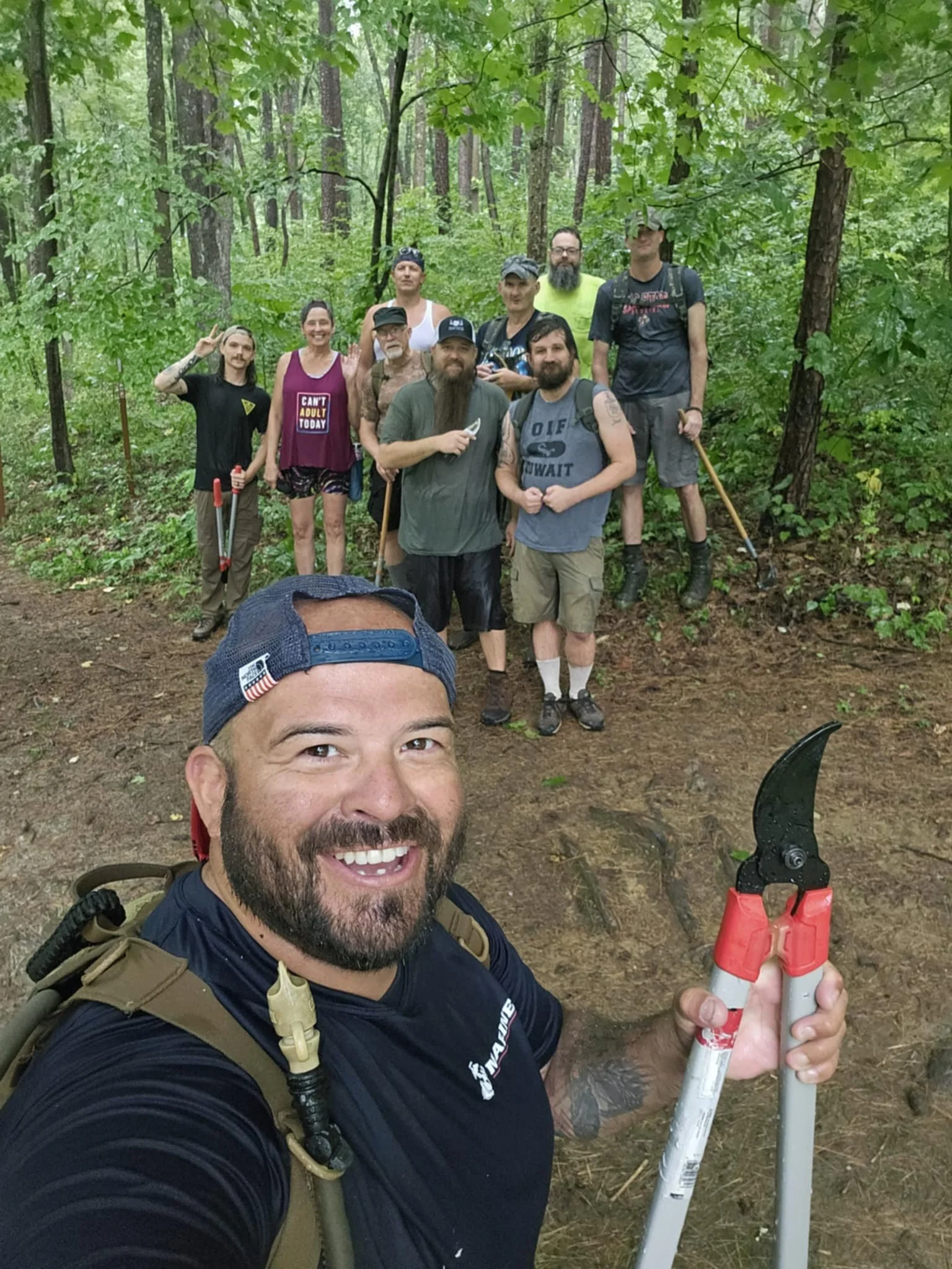 A group of people in a forest, some holding tools, and one person taking a selfie with a large tree root in the background.