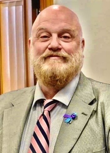 A middle-aged man with a bald head and a full, reddish beard wearing a gray suit, a white shirt, and a pink and navy striped tie. He has a blue ribbon pin on his suit lapel and is standing in front of a wooden door.