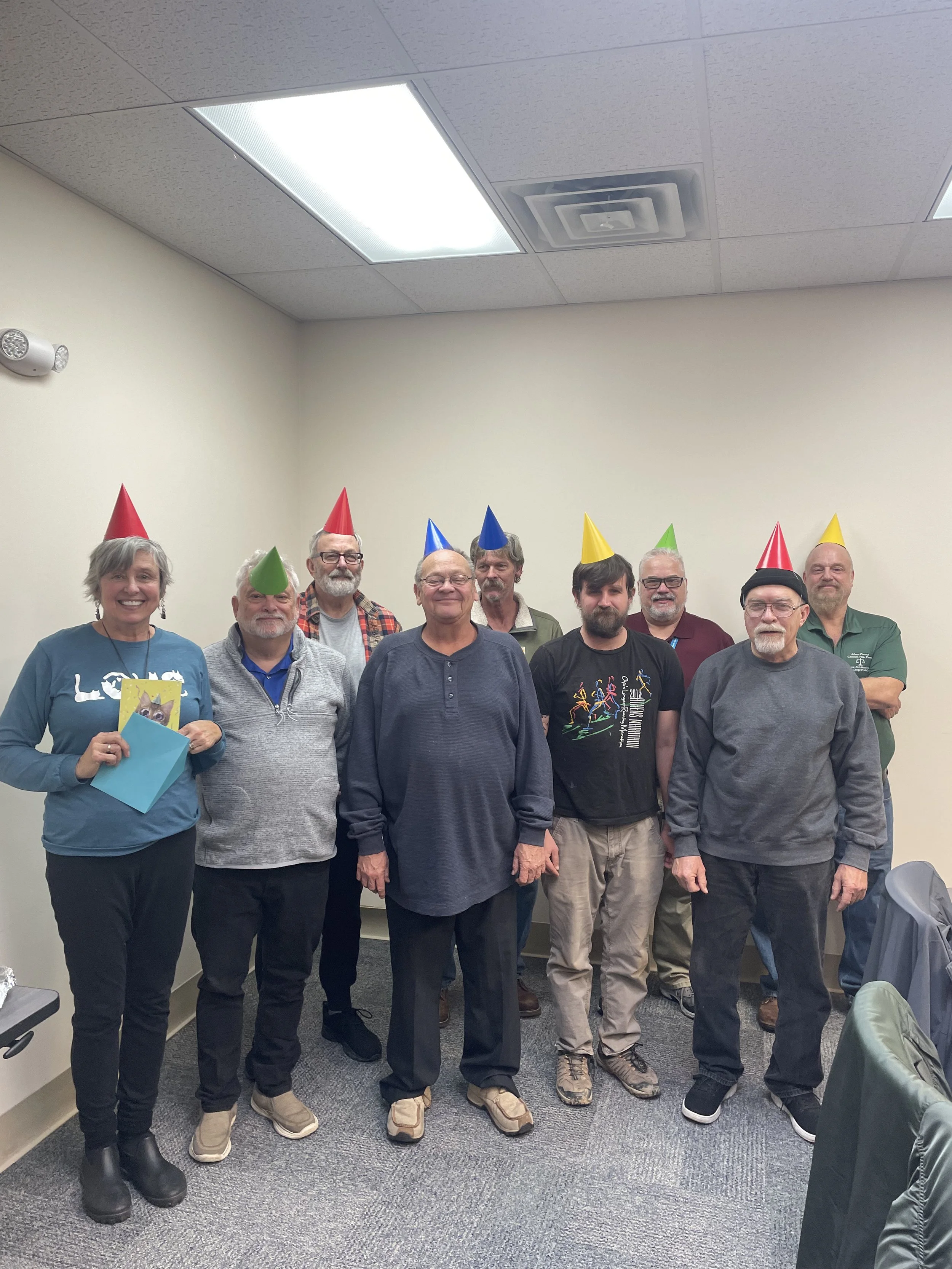 Group of ten adults in a room wearing colorful party hats, with some smiling and one holding a small gift or card