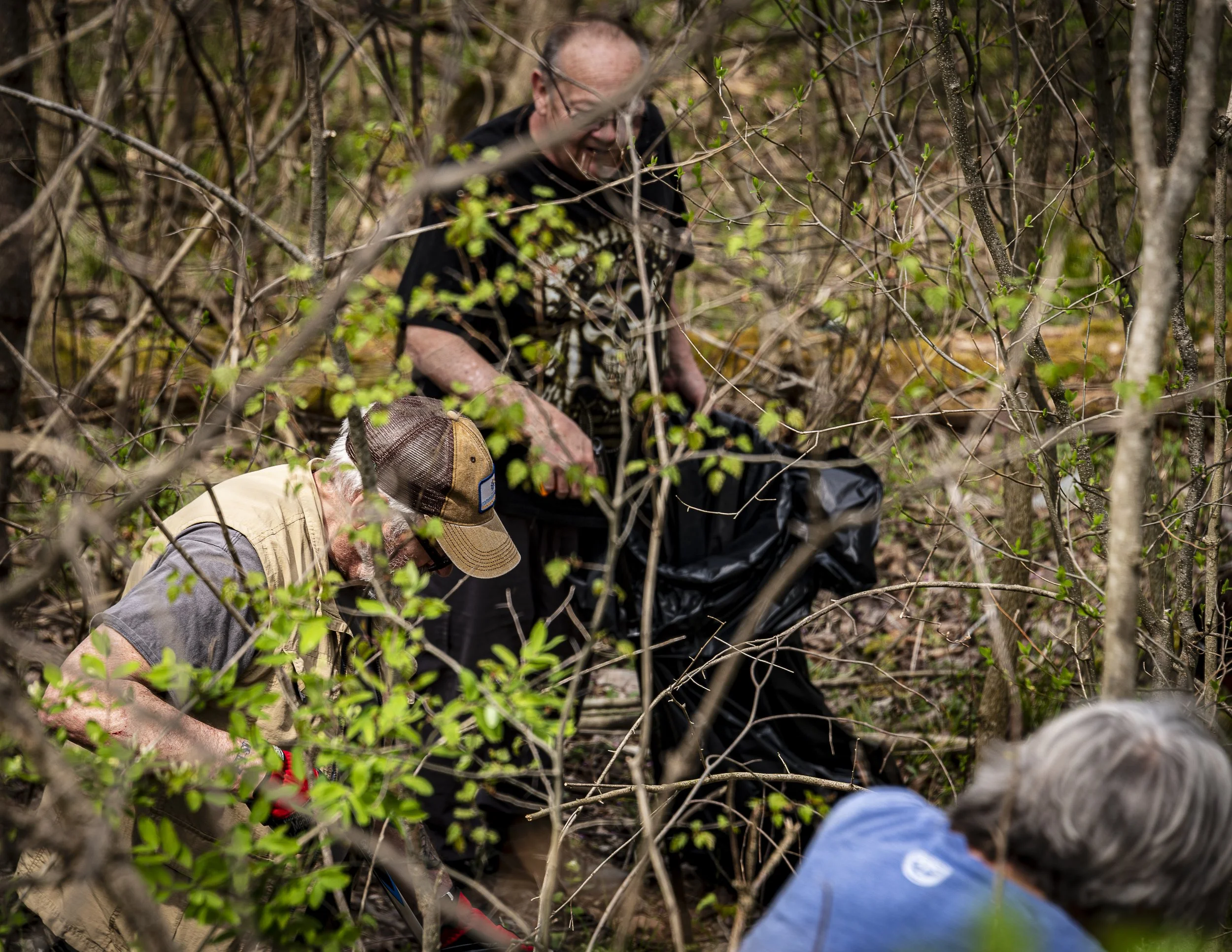 Three people are in a dense forest area surrounded by green leaves and thin branches. They appear to be exploring or working on some activity in the woods. One person is wearing a beige vest and cap, another is in a black shirt with glasses, and the 