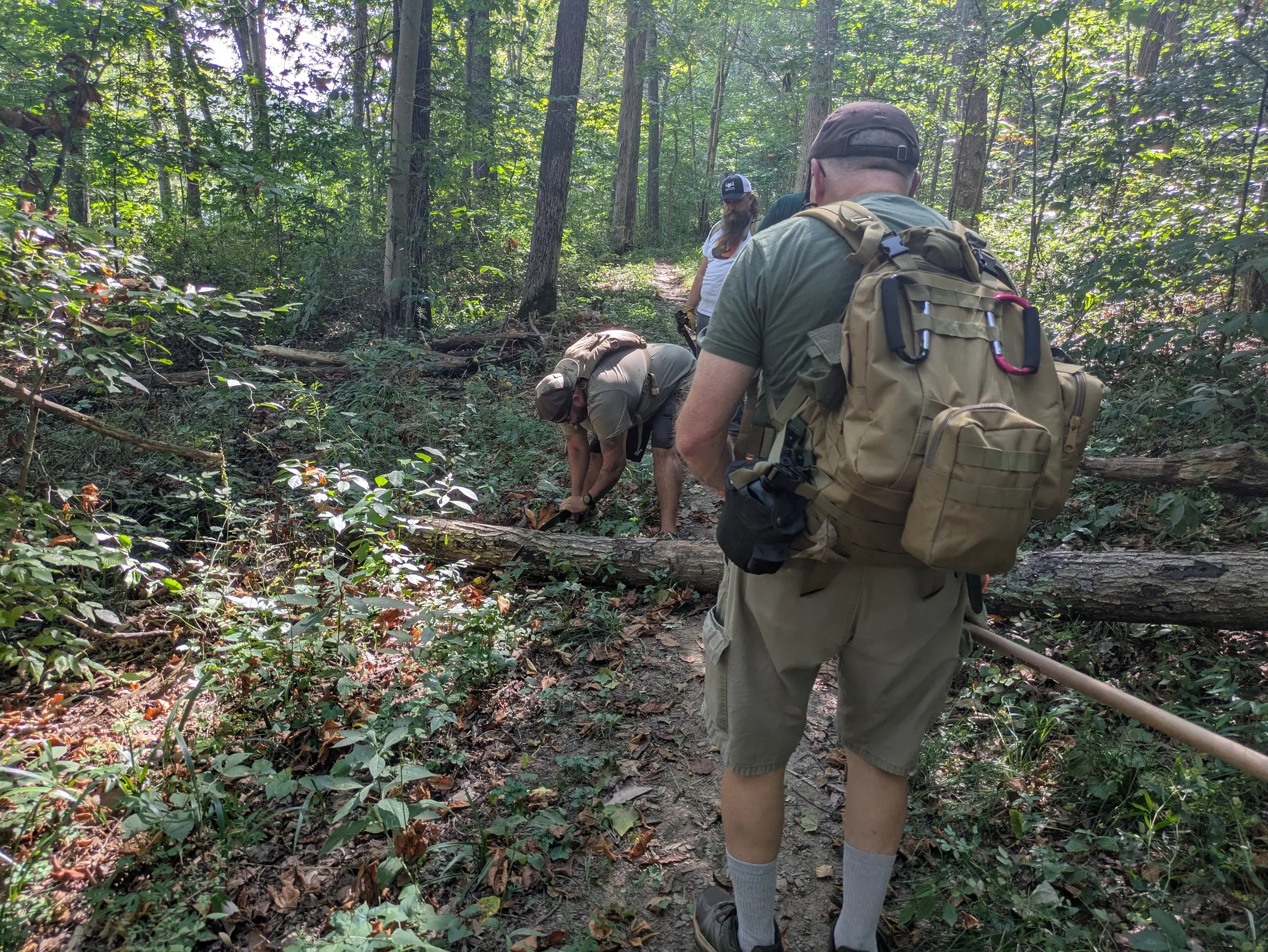 Group of people hiking in a dense forest, some bending down examining the ground, wearing casual outdoor clothing and backpacks.