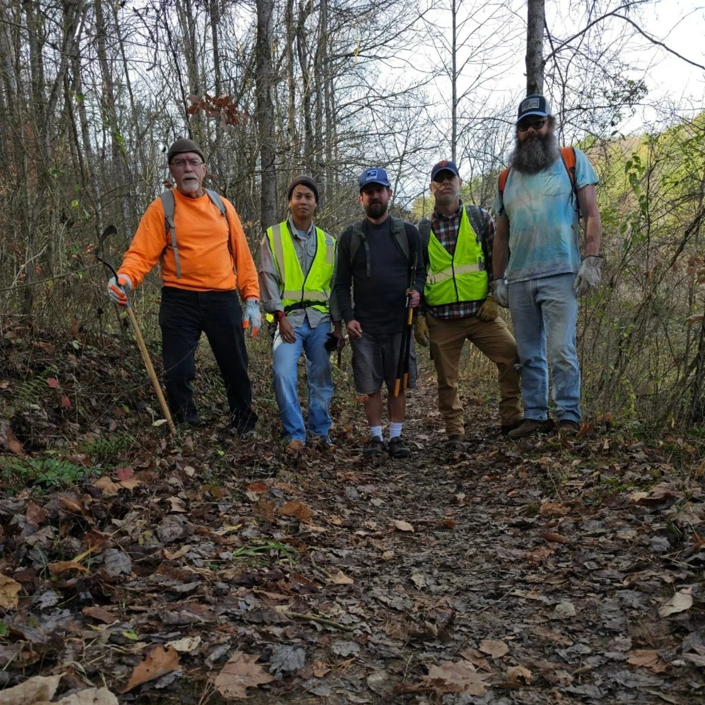 Five men standing on a wooded trail, wearing outdoor gear and reflective vests, indicating they are on a hike or outdoor project.
