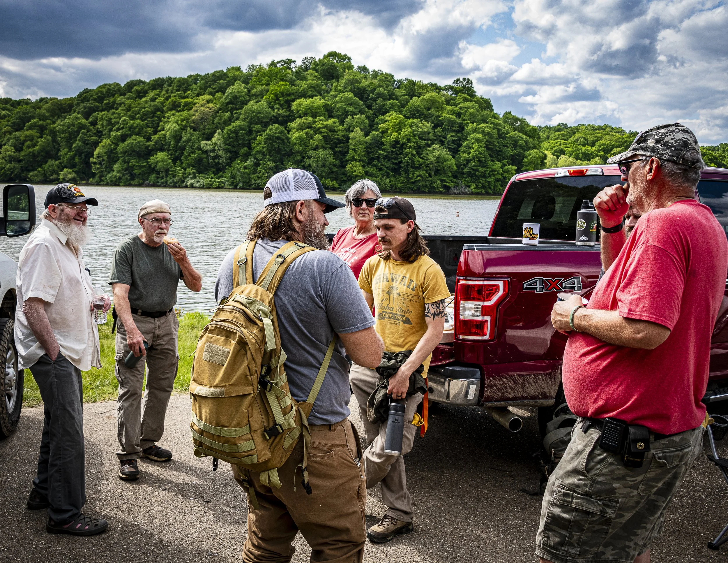 A group of people gathering outdoors near a water body with a forested hill in the background under a cloudy sky, standing beside a red truck.