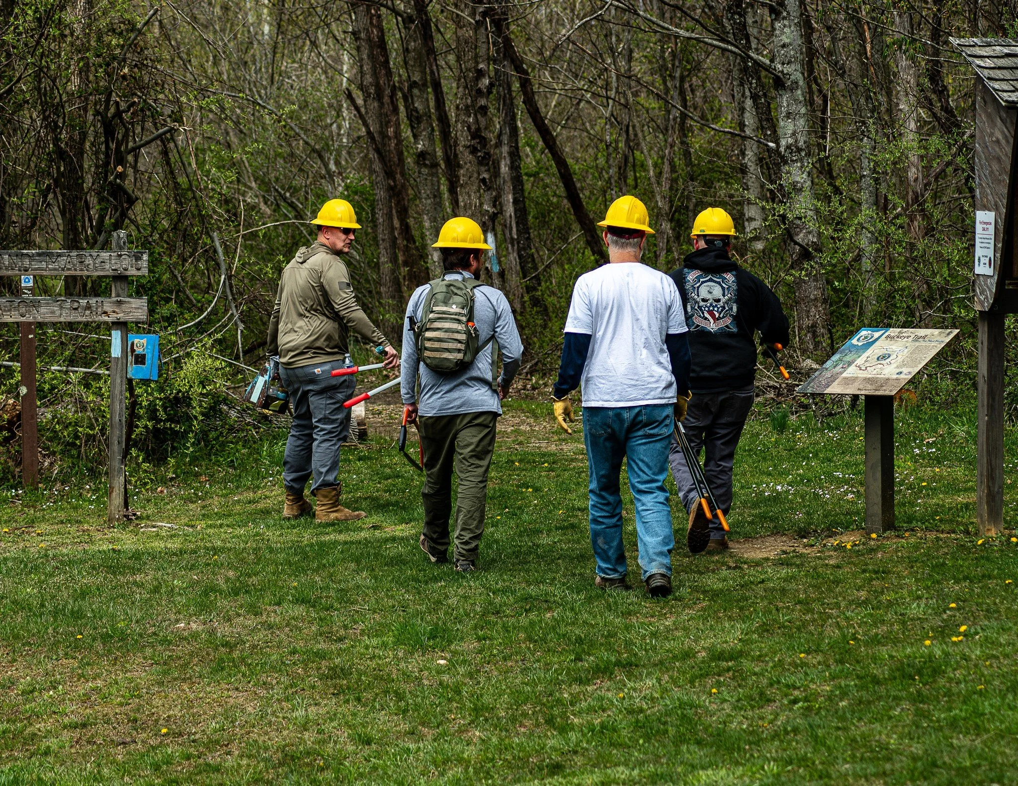 Group of five people wearing yellow safety helmets walking on a path through a wooded area with informational signs.
