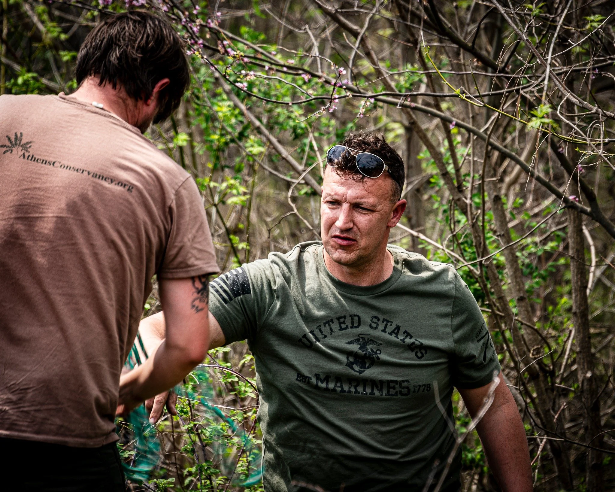 Two men are outdoors among tree branches and leaves, one with sunglasses on his head who appears to be talking or explaining something, and another man with his back turned facing the first. The man with sunglasses has short hair and a U.S. Marines T