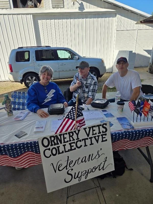 People sitting at a table with a sign for the Ornery Vets Collective, supporting veterans. The table has American flags and informational pamphlets. The setting appears to be outdoors, with a vehicle and building in the background.