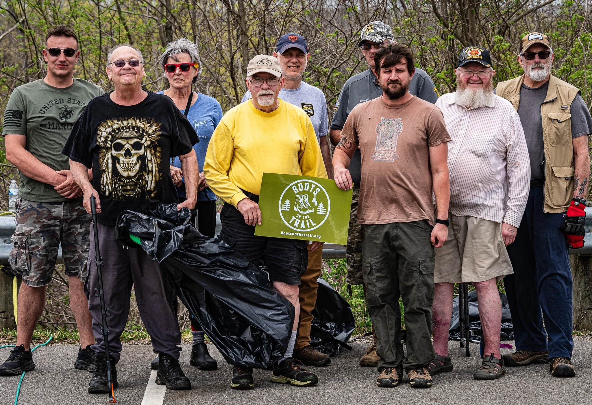 Group of people standing outdoors on a trail, holding a green sign that reads 'Boots on the Trail'. They are wearing casual and outdoor clothing, some with hats, sunglasses, and walking sticks, surrounded by trees and bushes.