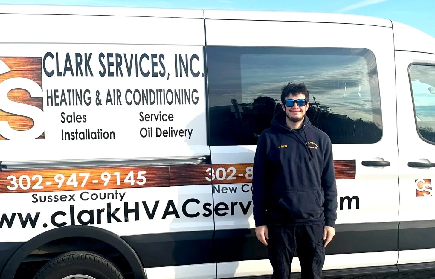 A man wearing sunglasses, a black cap, and a black hoodie with yellow text, standing in front of a sign.
