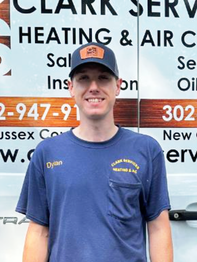 A young man in a blue work uniform and cap standing in front of a sign for Clark's Heating & Air Conditioning.