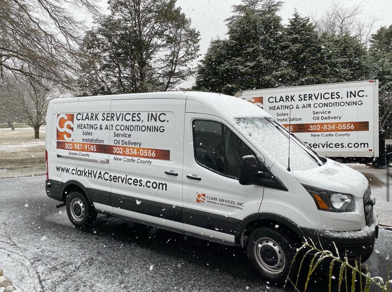 White service van with Clark Services Inc. branding, parked outdoors in a snowy setting with trees in the background. The van advertises heating and air conditioning services, including sales, installation, service, and oil delivery, with contact numbers and website.