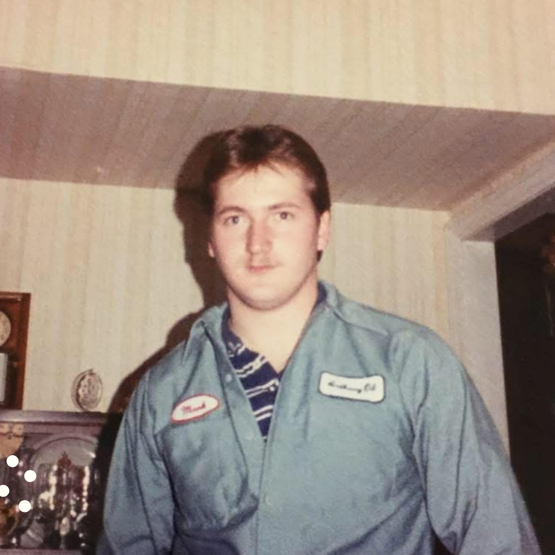 A young man with brown hair and a light complexion, wearing a light gray work uniform with red and white name patches, standing indoors with a striped wallpaper background.