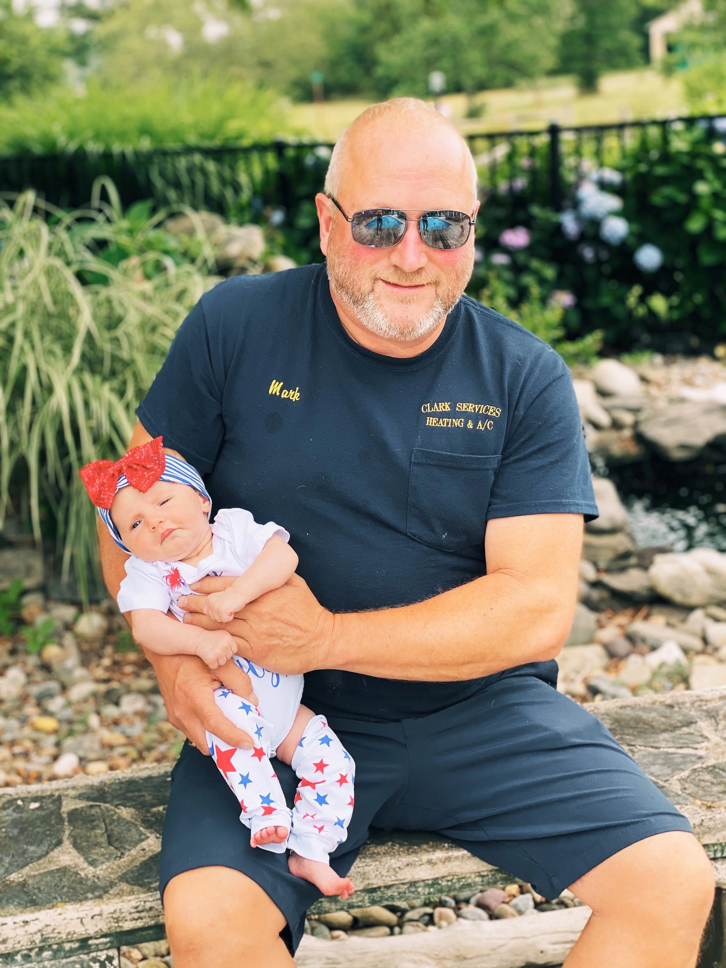 A man wearing sunglasses, a navy blue t-shirt with yellow embroidery, and black shorts holds a small baby dressed in patriotic-themed clothing with stars and stripes, sitting on a stone bench outdoors in a garden setting.