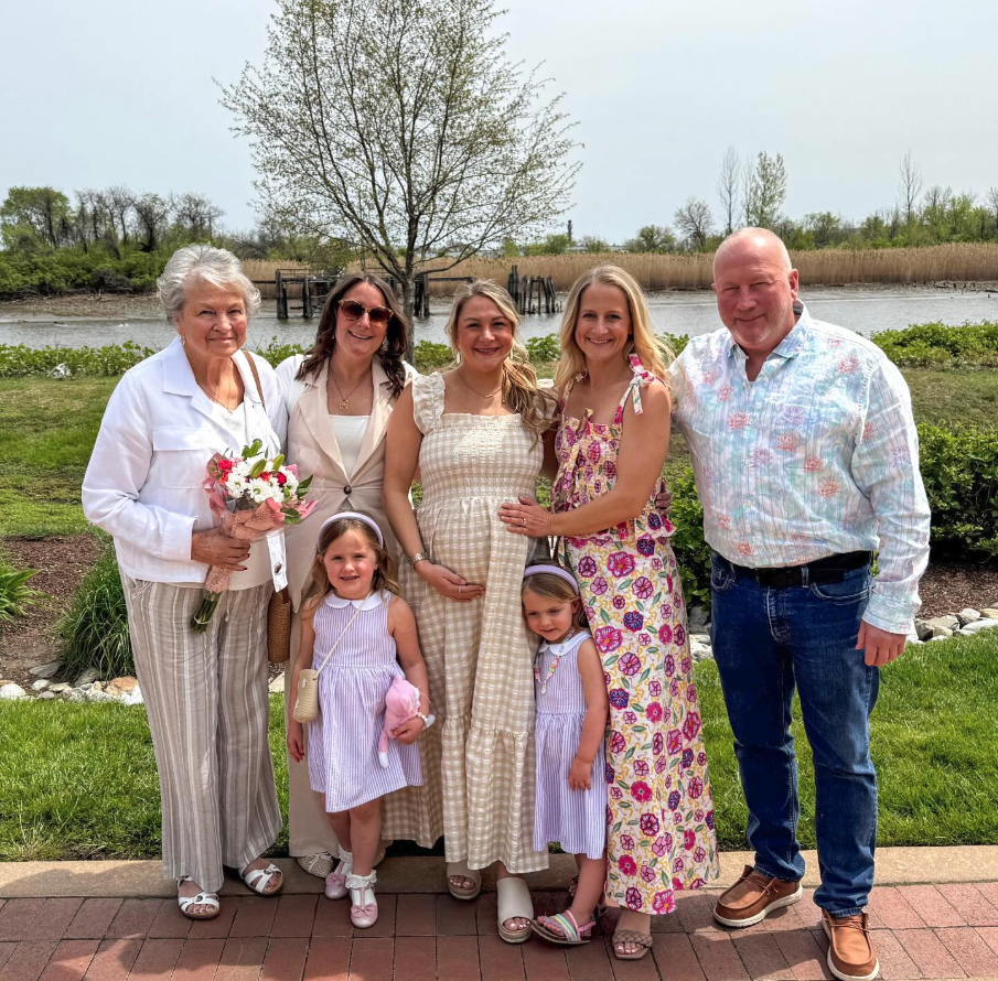 Family group outdoors near a river with trees in the background, consisting of an elderly woman holding a bouquet, two young girls in purple dresses, three women, and a man smiling at the camera.