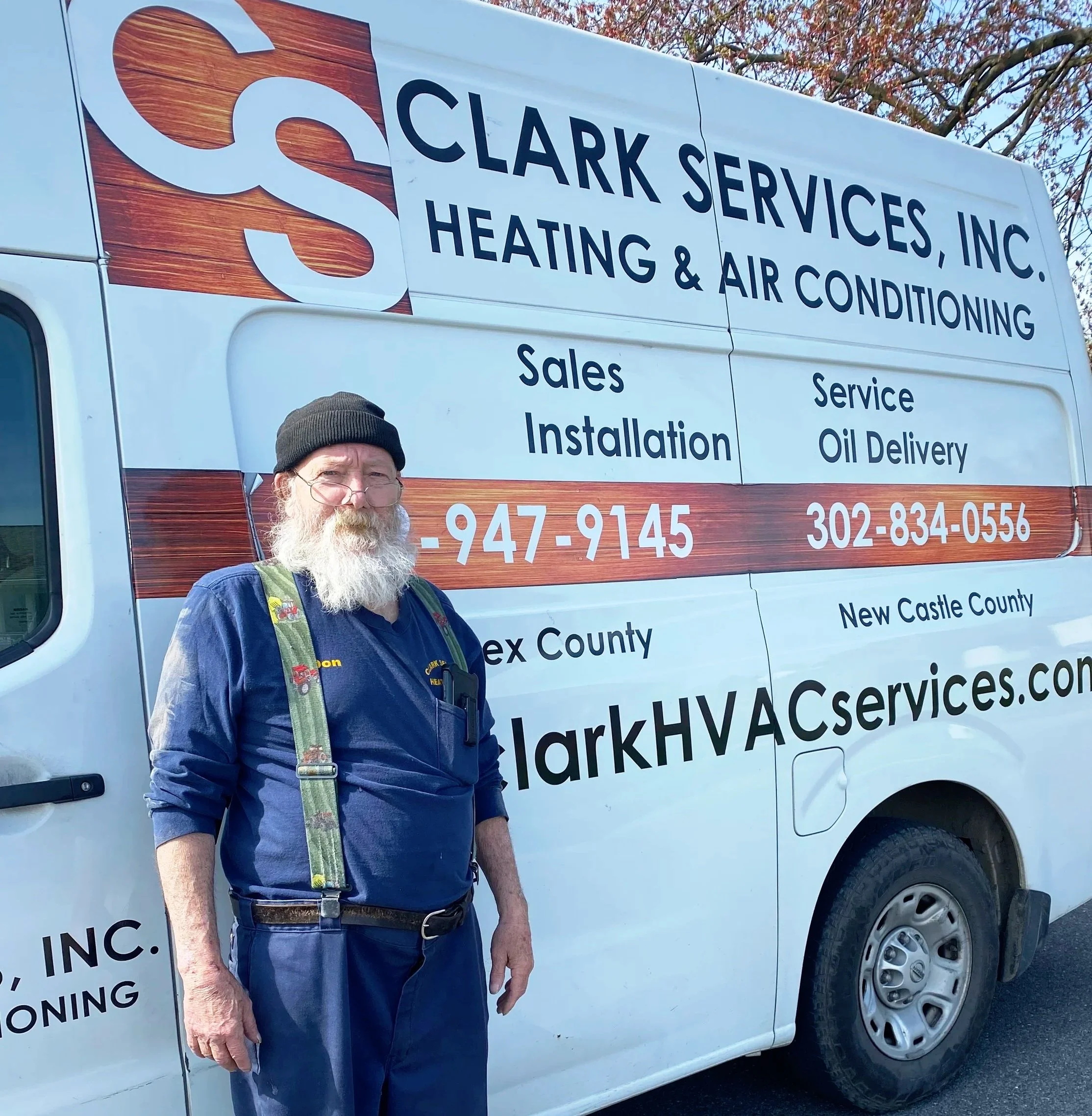 Man in blue overalls and black cap standing next to Clark Services van with heating and air conditioning advertisement.