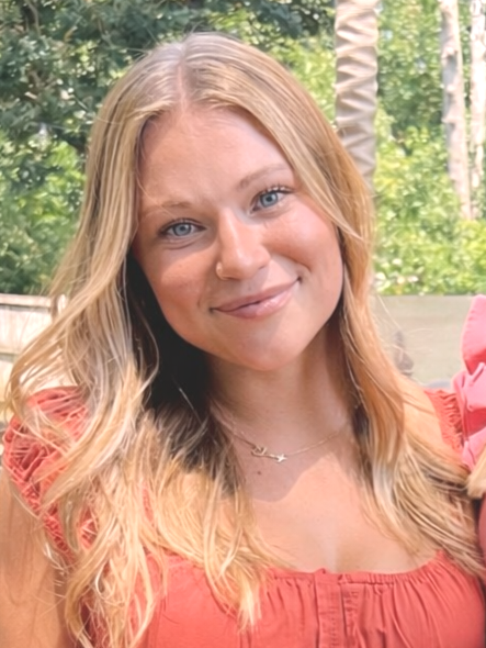 A young woman with long blonde hair smiling outdoors beside a pond with lily pads and surrounded by trees.