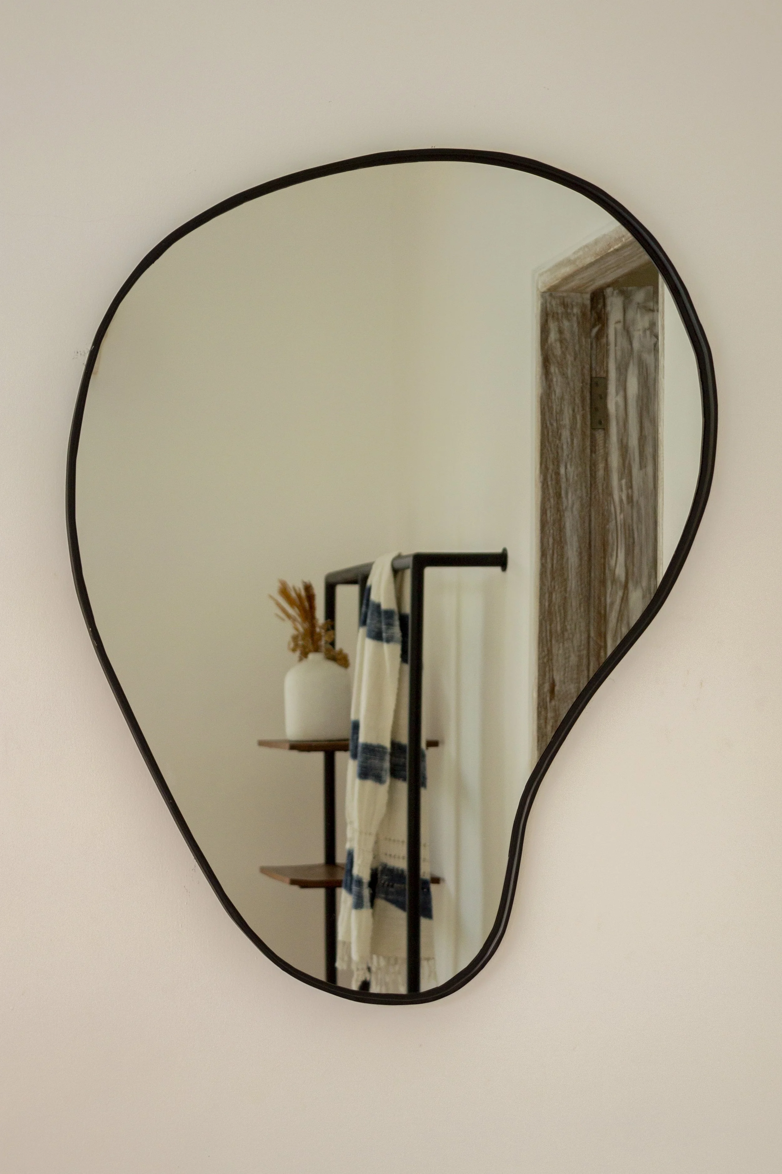 A heart-shaped mirror on a wall reflecting a wooden shelf with a white vase containing dried flowers and a ladder with a striped blanket draped over it.
