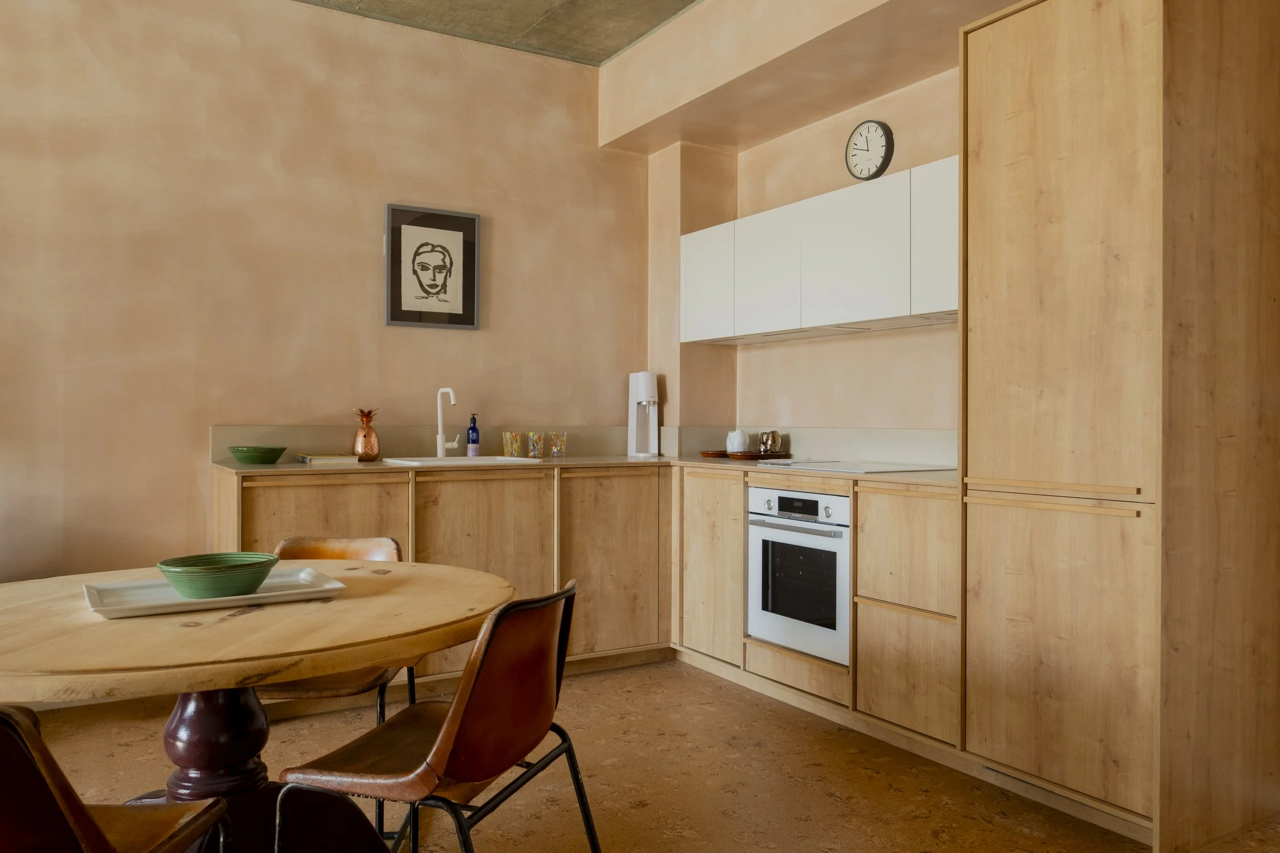 A kitchen with wooden cabinets, a round wooden dining table with green bowls and plates, a retro-style chair, a wall clock, framed artwork, and a framed photo of a woman's face