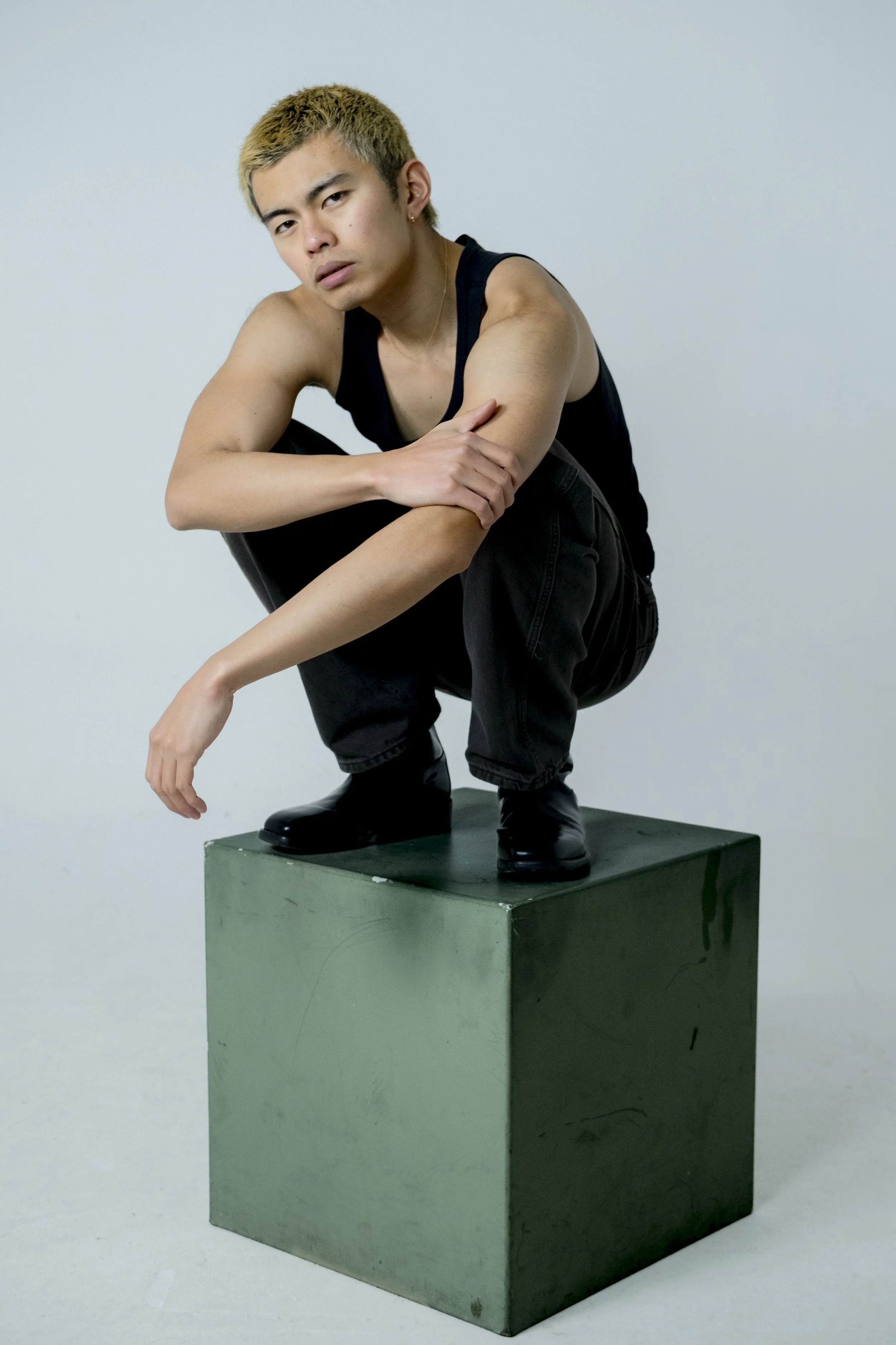 A young man with bleached blonde hair wearing a black tank top and black pants, squatting on a green wooden block against a plain light gray background.