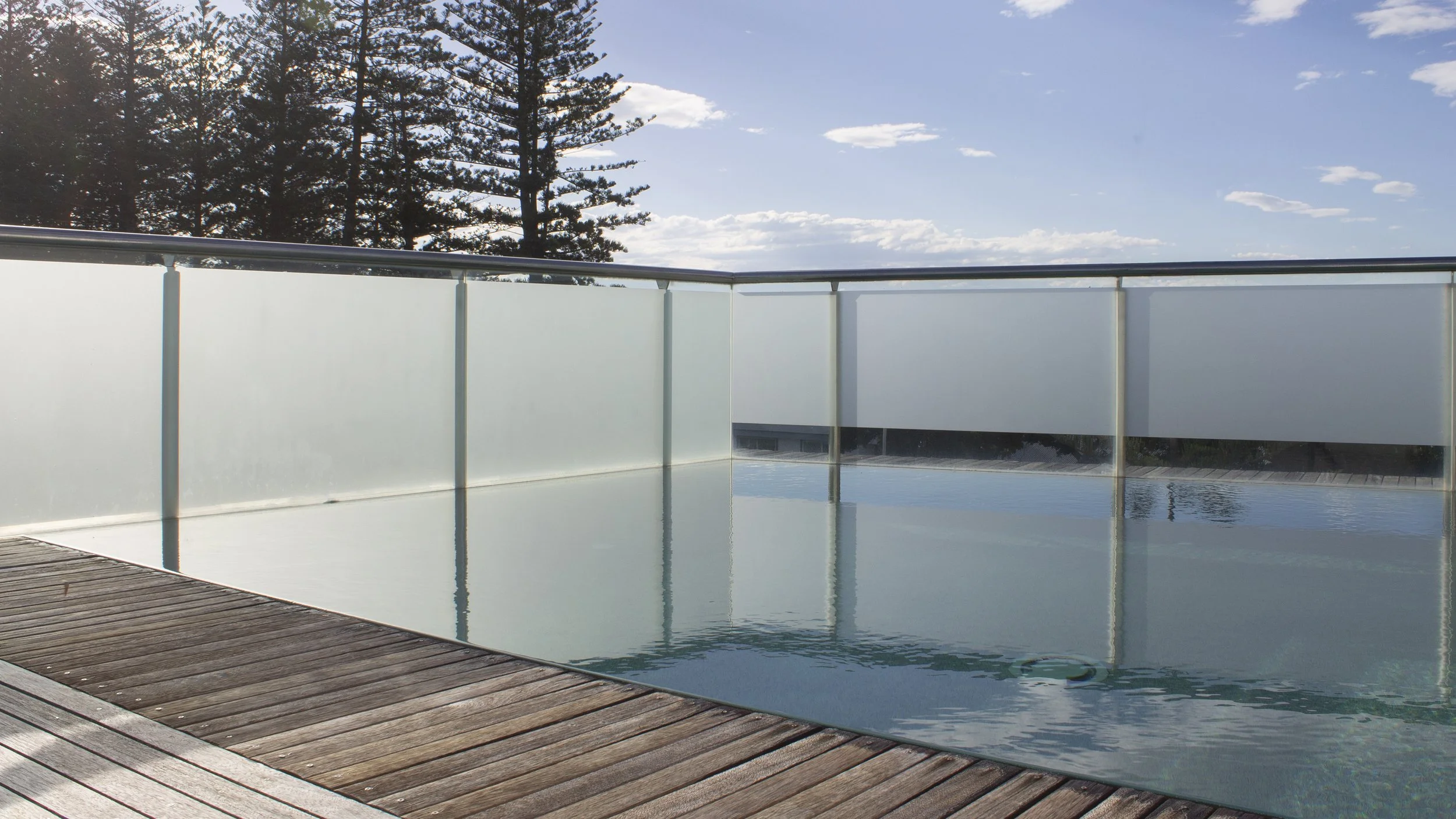 A swimming pool with a wooden deck and a glass barrier, with trees and a blue sky with clouds in the background.