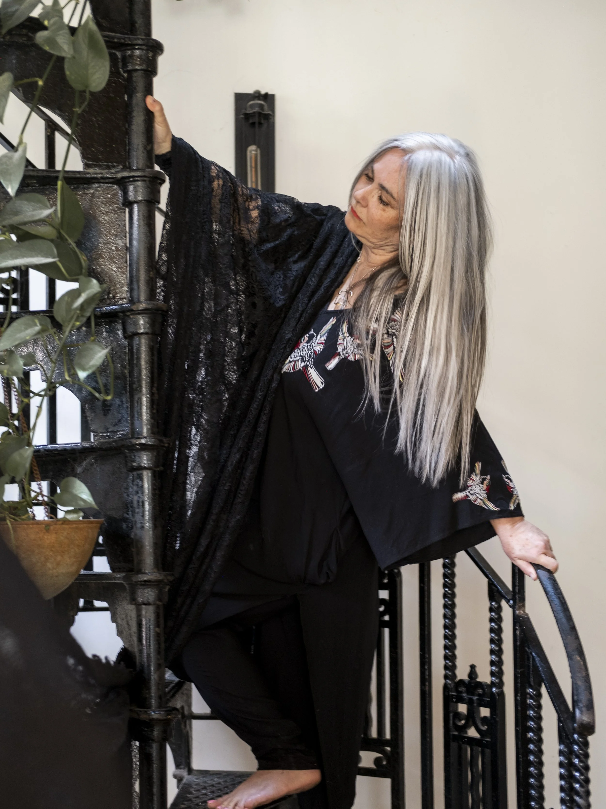 An elderly woman with long gray hair adjusting a staircase with a black lace shawl over her shoulders. She is wearing a black dress with embroidered hummingbirds on the front and has a calm expression.