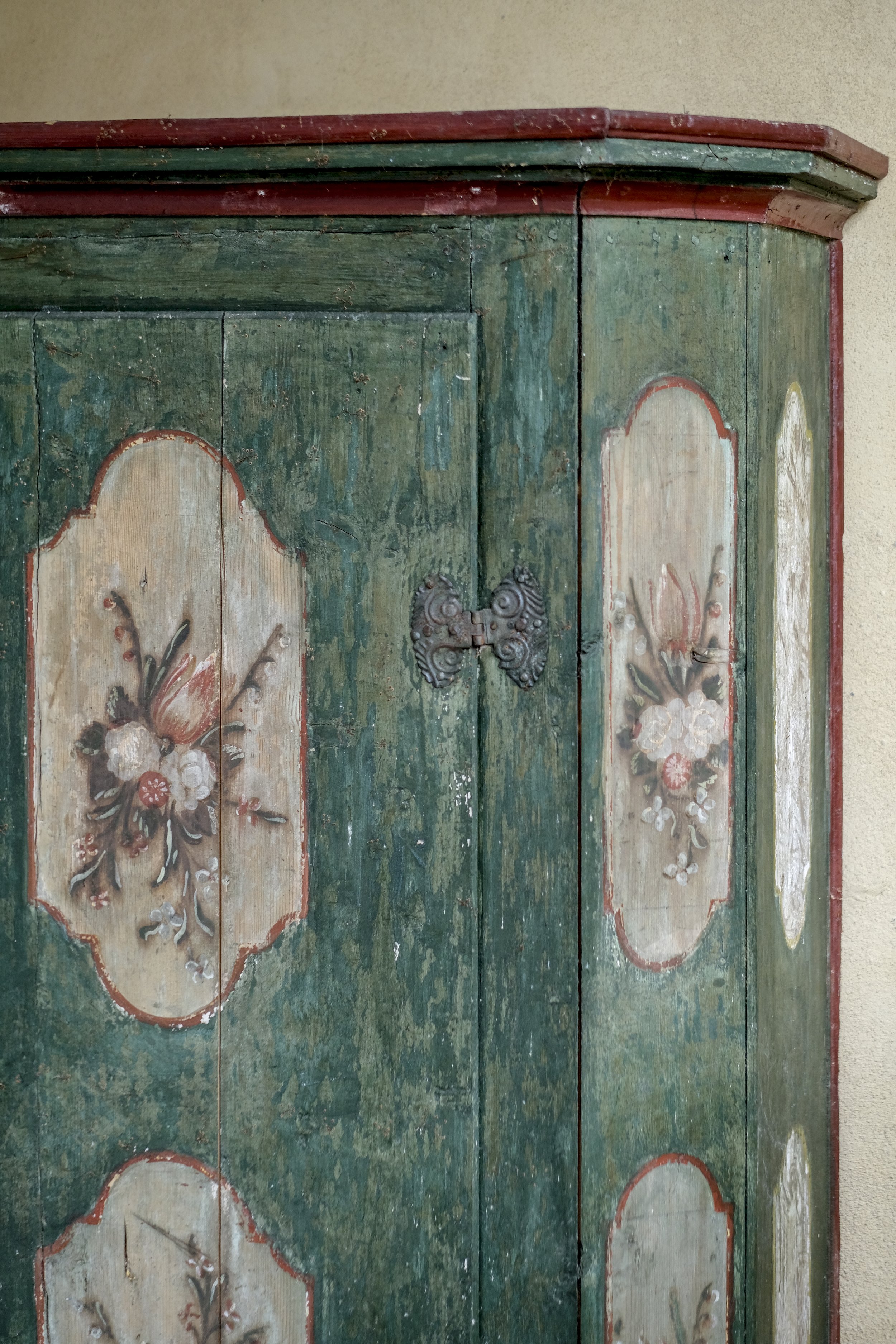Close-up of an antique wooden cabinet with faded green paint, floral painted panels, and ornate metal hinges, against a beige wall.
