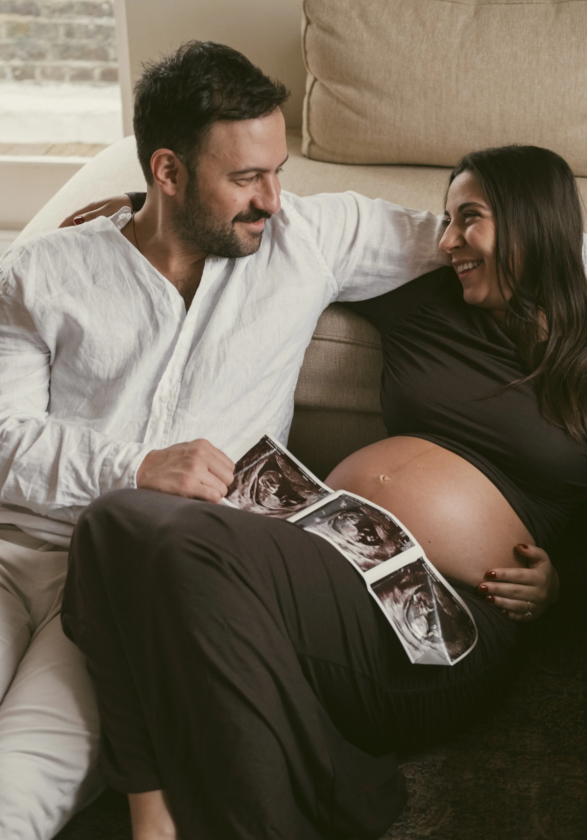 A pregnant woman and a man sitting on the floor, smiling at each other, with ultrasound images of the pregnancy on her belly. maternity couples photoshoot