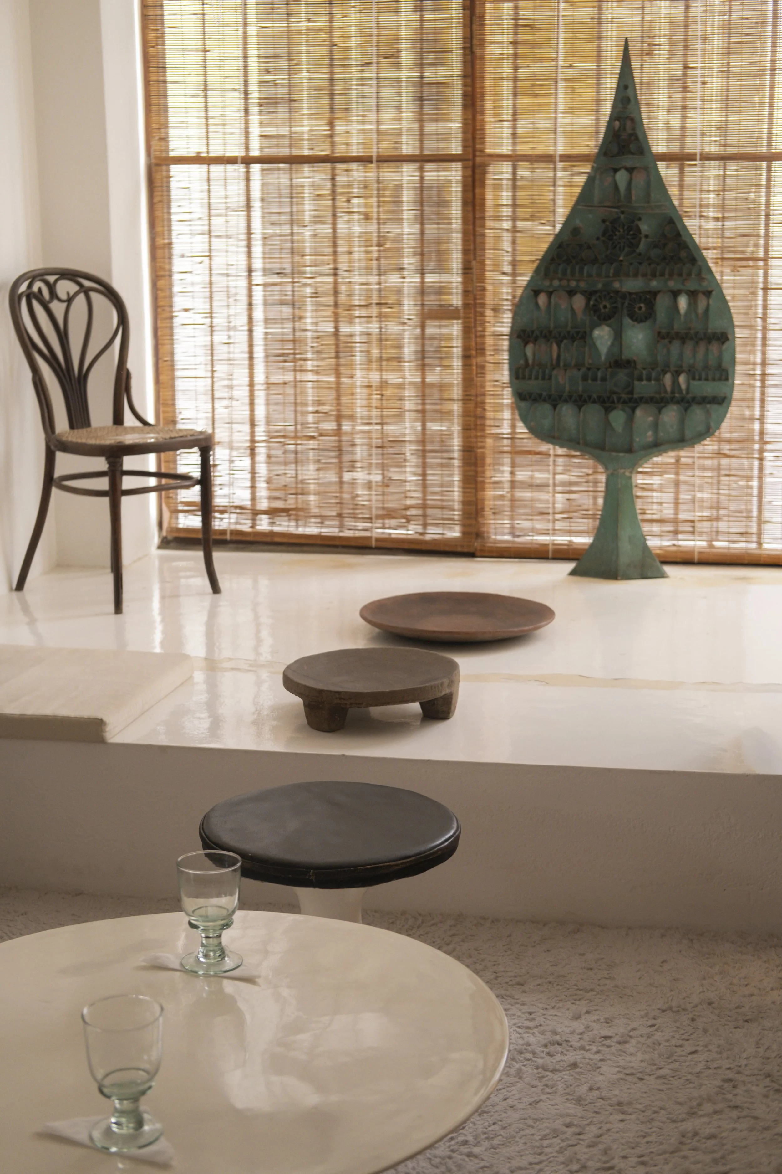 Interior of a room with a white round table, a glass on it, black stools, a wooden chair, and a decorative green sculpture on a stand in front of bamboo blinds covering windows.