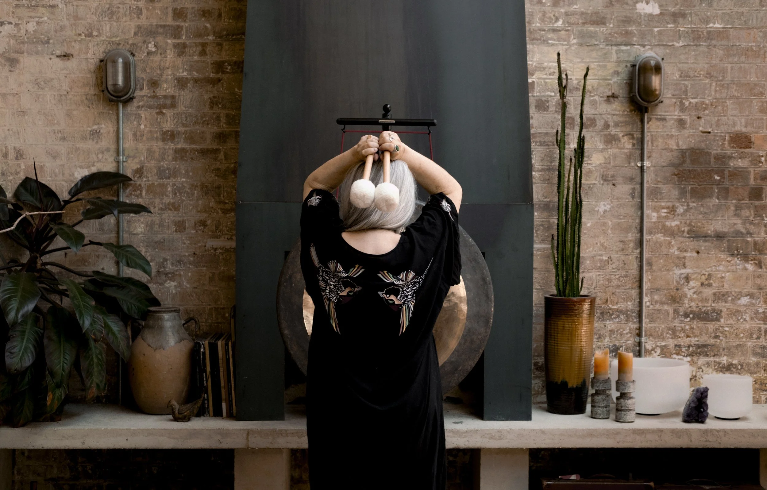 A person with gray hair is holding a mallet in front of their face, with their arms raised above their head, in a room with a brick wall and various decorative items including potted plants, candles, and a large gong.