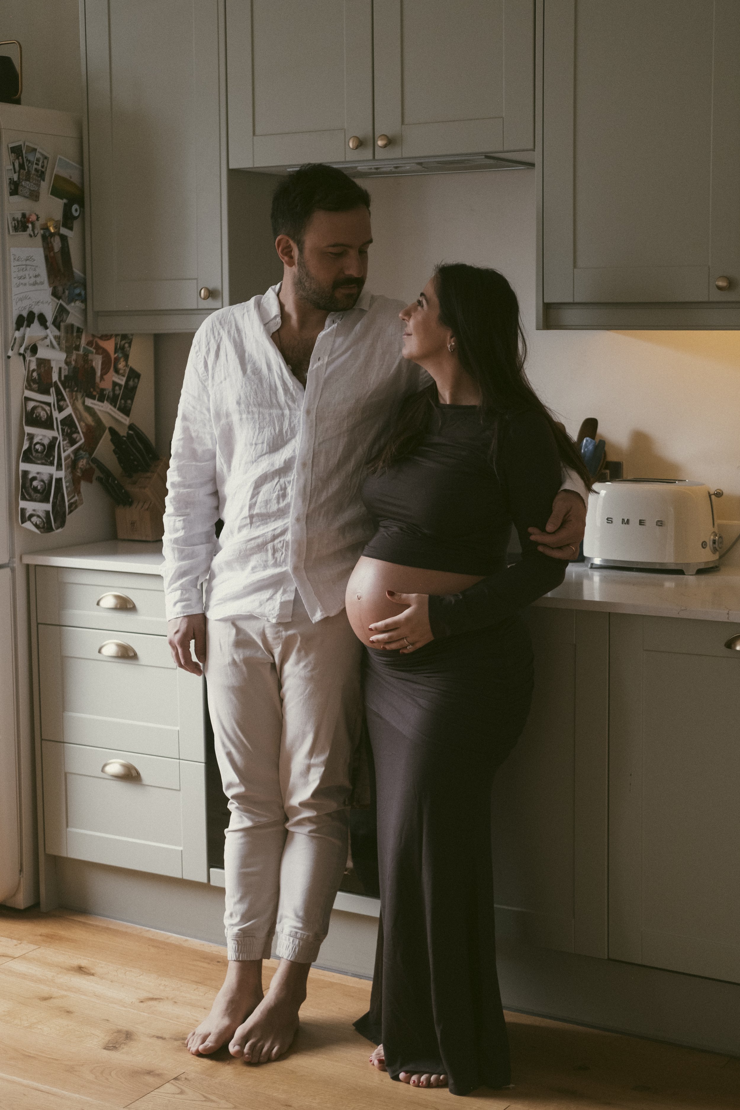 A pregnant woman and a man standing in a kitchen, smiling and looking at each other, with the woman holding her belly. The man is wearing a white shirt and beige pants, and the woman is wearing a black long sleeve top and dark pants.