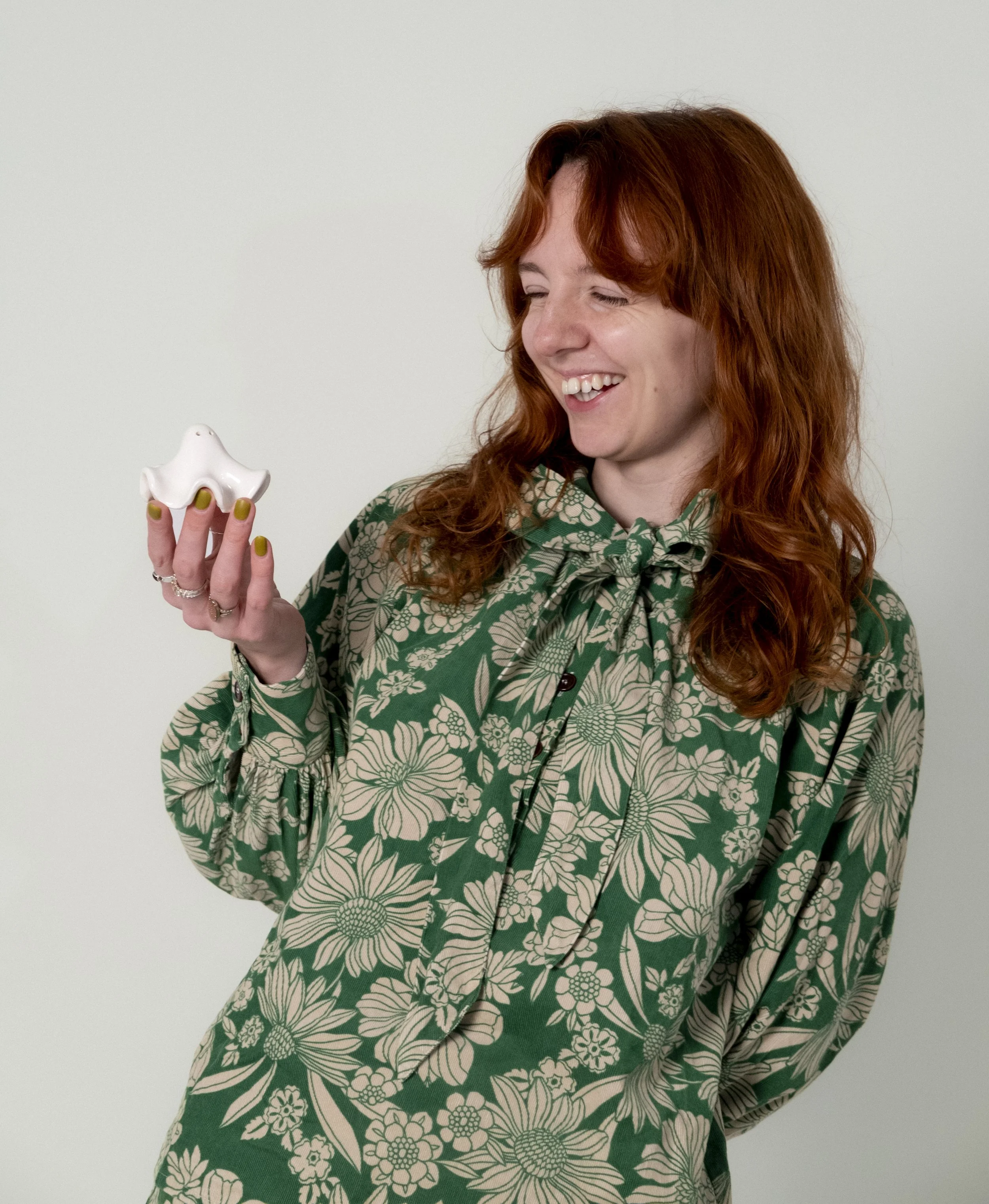 Woman with red hair smiling while holding a white porcelain sculpture of an abstract face, wearing a green floral patterned shirt, standing against a plain white background.