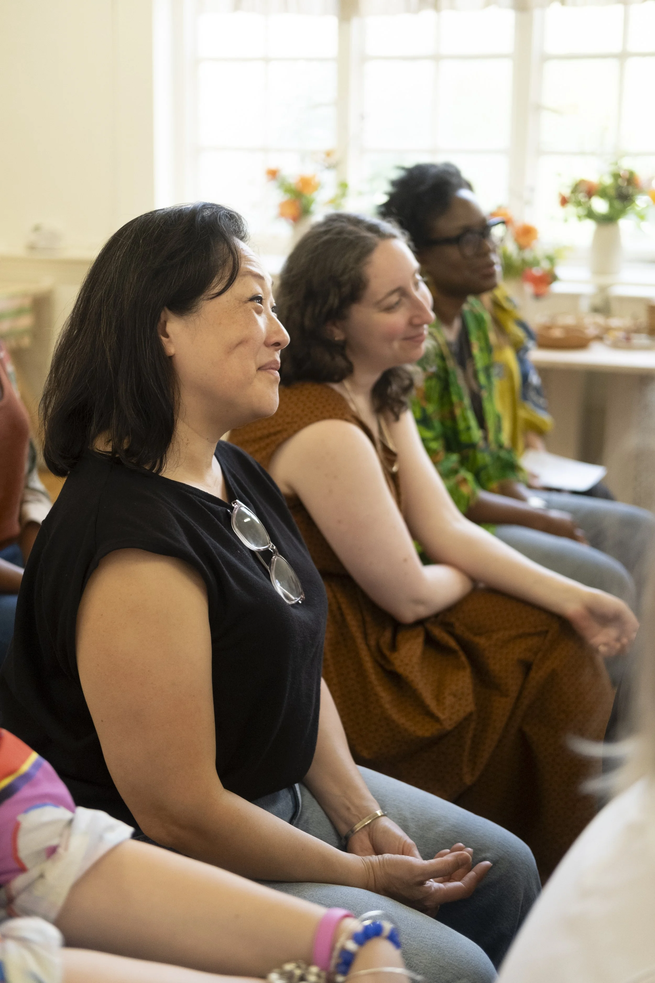 Three women sitting in a row, attentively listening in a well-lit room with large windows and flower arrangements.
