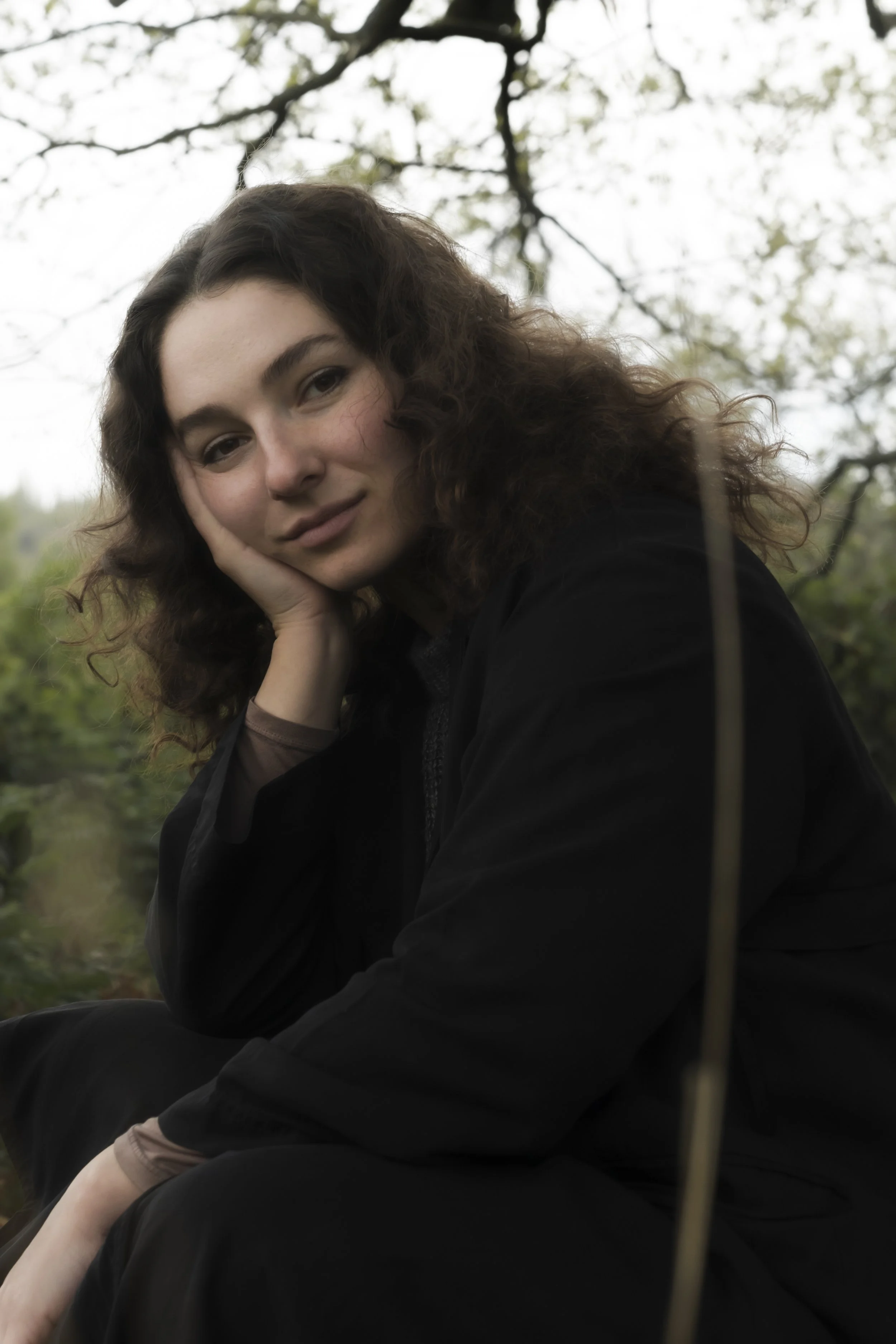 A young woman with curly brown hair resting her face on her hand outdoors during daytime, with a background of tree branches and open sky.