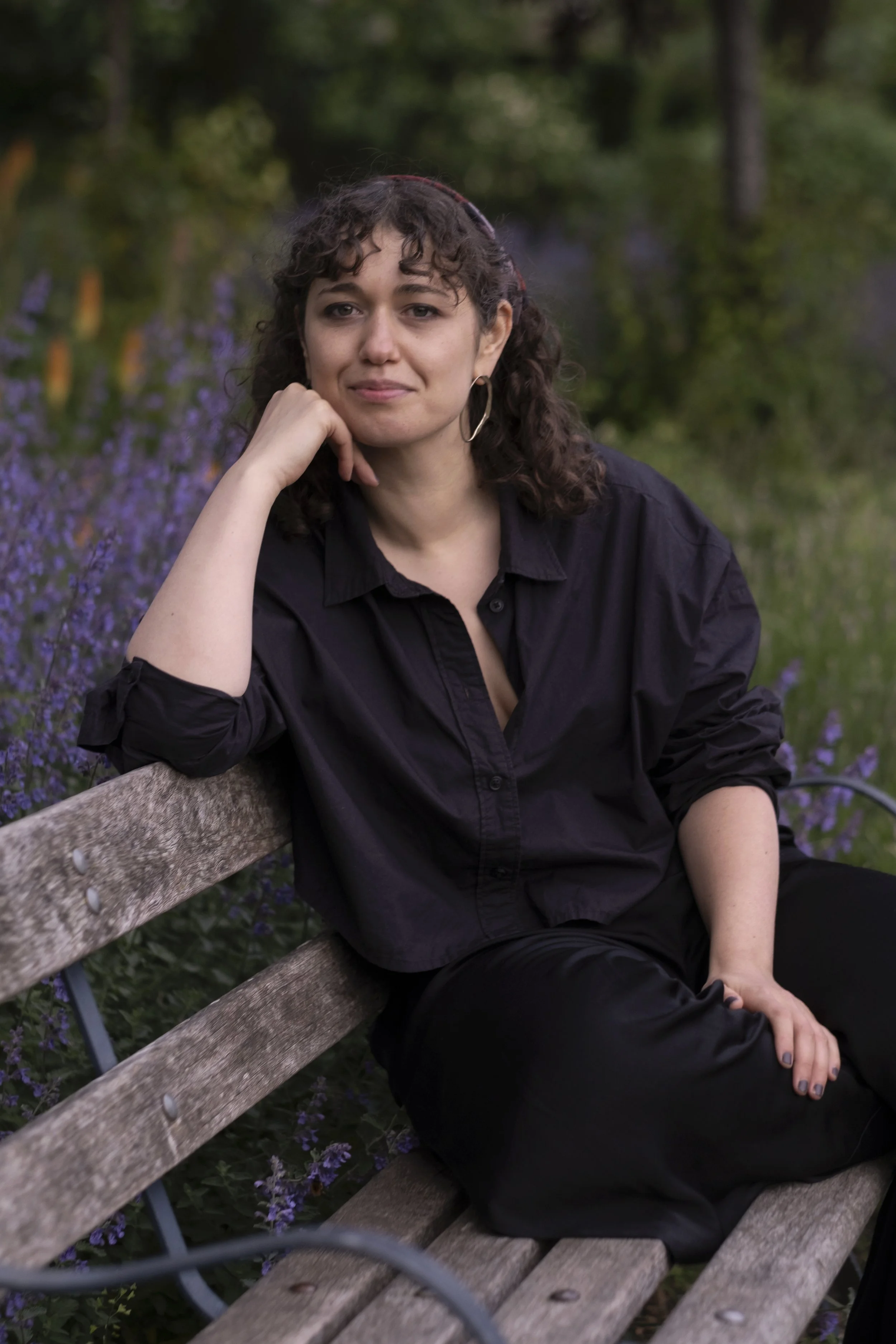 A woman with curly dark hair, wearing a black shirt and black pants, sitting on a wooden park bench in a garden with purple flowers and green trees in the background.